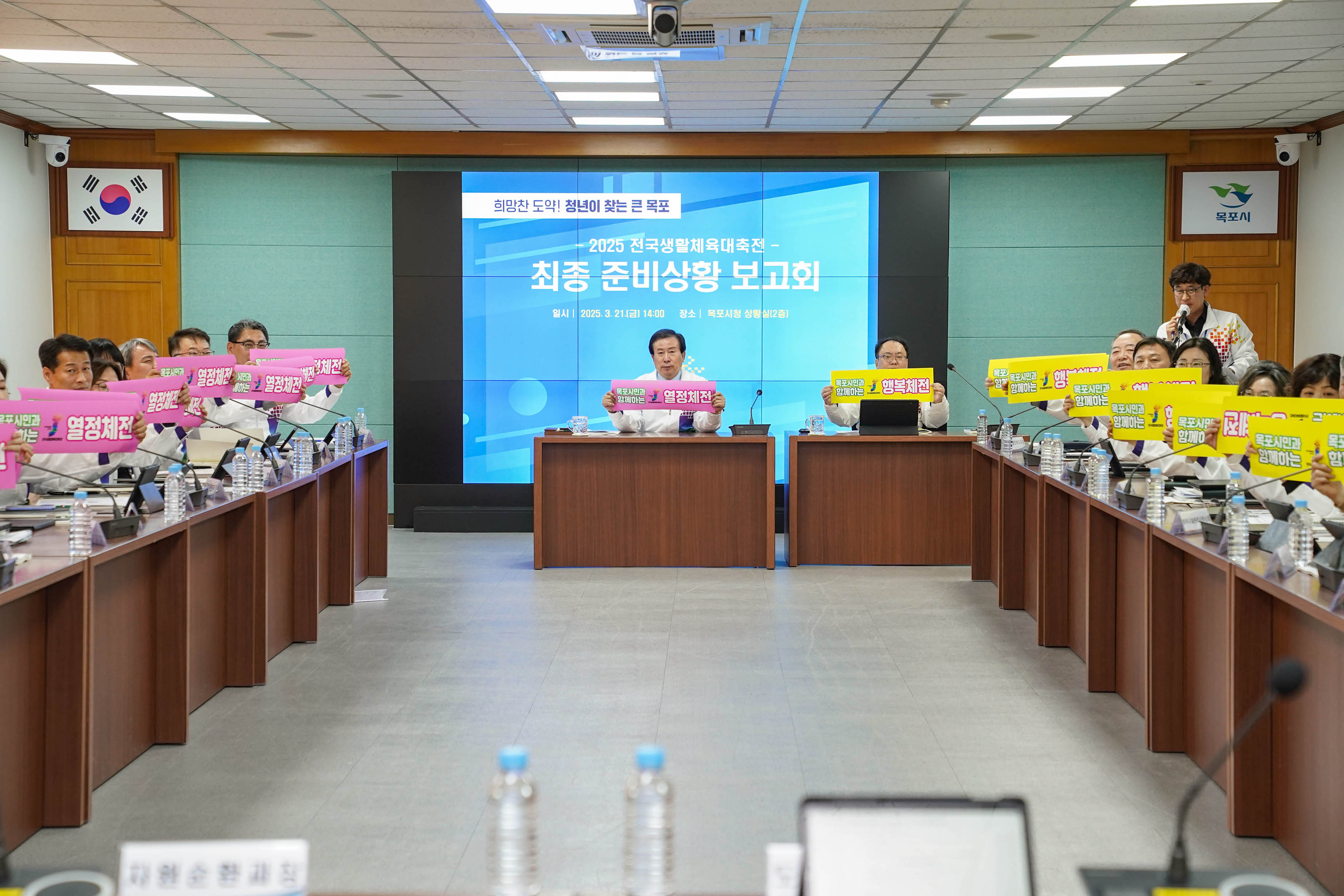 '희망찬 도약! 청년이 찾는 큰 목포 2025 전국생활체육대축전 - 최종 준비상황 보고회' 문구가 적힌 스크린 앞으로 박홍률 목포시장을 비롯한 참가자들이 배너를 들고 있는 모습. 배너에는 '열정체전', '행복체전' 이라는 문구가 적혀있음