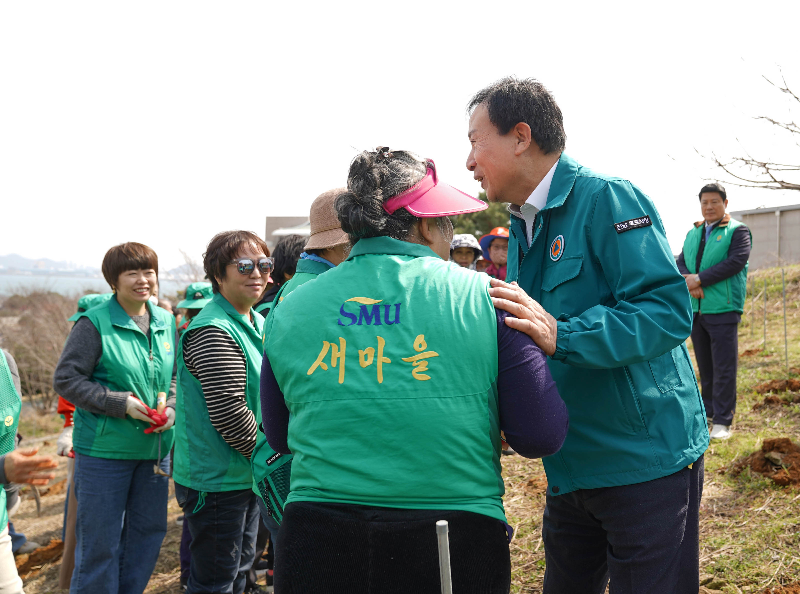 'SMU 새마을'이라고 적힌 초록색 조끼를 입고 자원봉사자들이 함께 활동하는 모습. 박홍률 목포시장이 다른 사람과 악수를 나누고 있으며, 다른 자원봉사자들이 주변에서 서 있는 모습