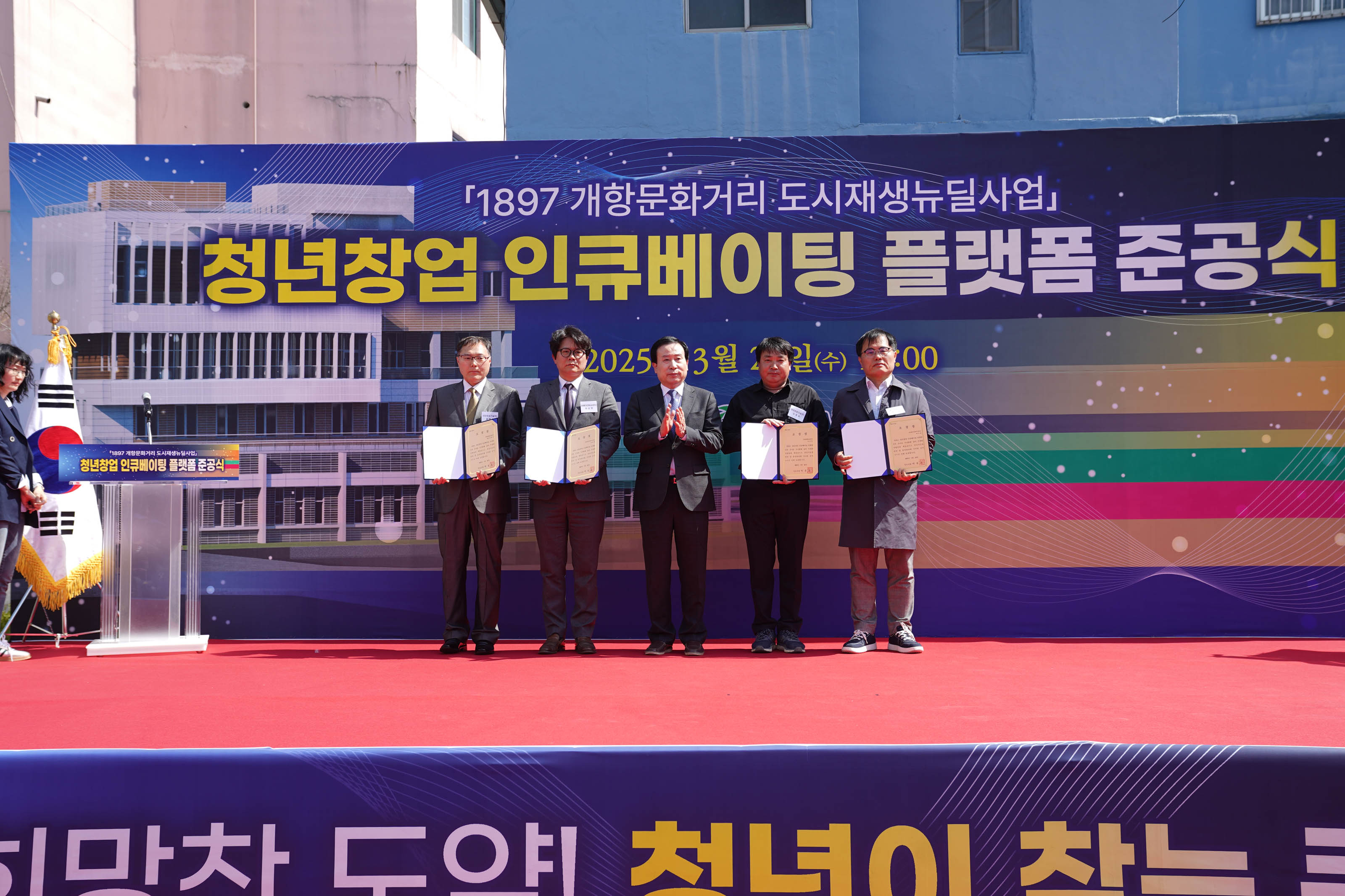 2025년 3월 26일, 목포시에서 열린 ‘1897 개항문화거리 도시재생뉴딜사업 - 청년창업 인큐베이팅 플랫폼 준공식’ 행사 현장. 무대 위에는 양복을 입은 박홍률 목포시장과 4명의 관계자들이 서 있으며, 그중 4명이 임명장 또는 인증서를 펼쳐 보이고 있고, 시장은 박수를 치고 있다. 배경에는 준공식 타이틀과 함께 '청년창업 인큐베이팅 플랫폼 준공식', '2025년 3월 26일(수) 14:00'이라는 글씨가 적혀 있다.