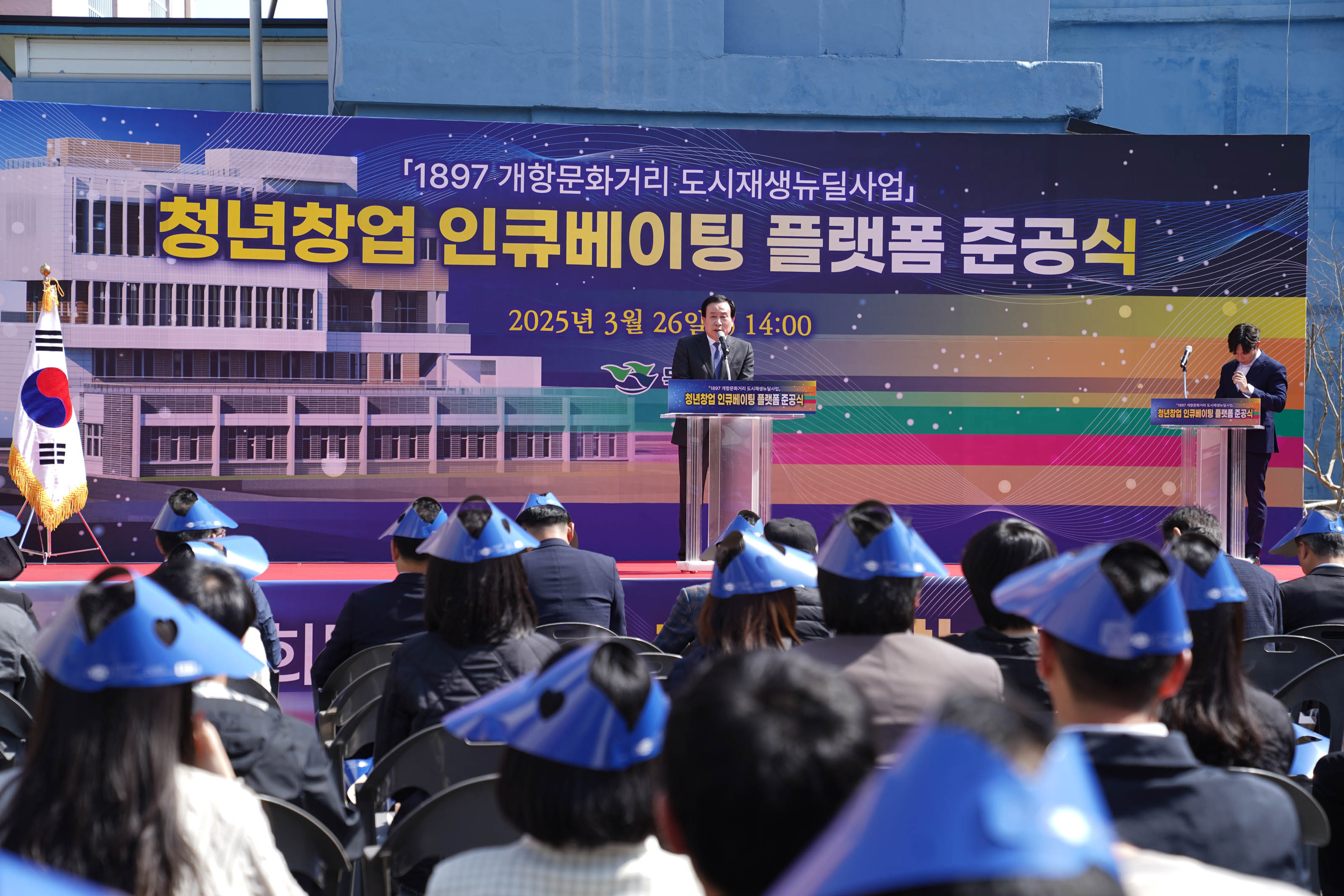 2025년 3월 26일 오후 2시, 목포시에서 열린 ‘청년창업 인큐베이팅 플랫폼 준공식’ 행사 모습. 무대 위 연단에서 정장을 입은 박홍률 목포시장이 마이크를 들고 발언 중이며, 좌측 태극기와 우측 사회자가 보인다. 청중은 파란색 종이모자(햇빛가리개 모자)를 착용한 상태로 정면 무대를 바라보고 있다. 배경에는 '1897 개항문화거리 도시재생뉴딜사업 청년창업 인큐베이팅 플랫폼 준공식'이라는 문구와 목포시 로고가 보인다.