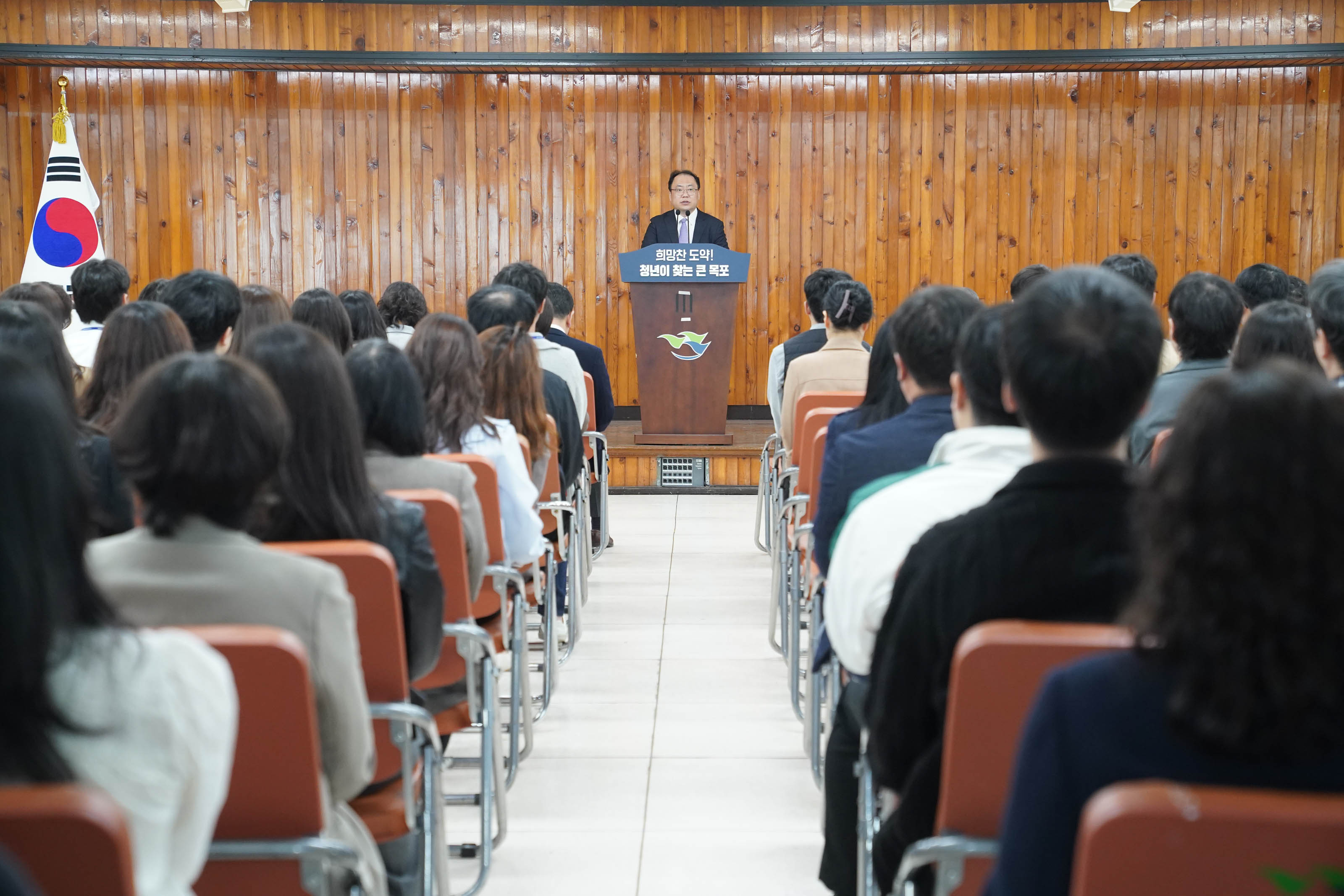 나무 벽면이 있는 강당에서 진행된 특강 현장. 중앙 단상에 한 인사가 서 있으며, 단상에는 '희망찬 도약! 청년이 찾는 큰 목포'라는 문구가 적혀 있다. 단상 앞에는 목포시 로고가 있고, 왼쪽에는 태극기가 세워져 있다. 청중으로 보이는 청년 및 관계자들이 단상을 향해 앉아 있으며, 모두 정면을 주시하고 있다.