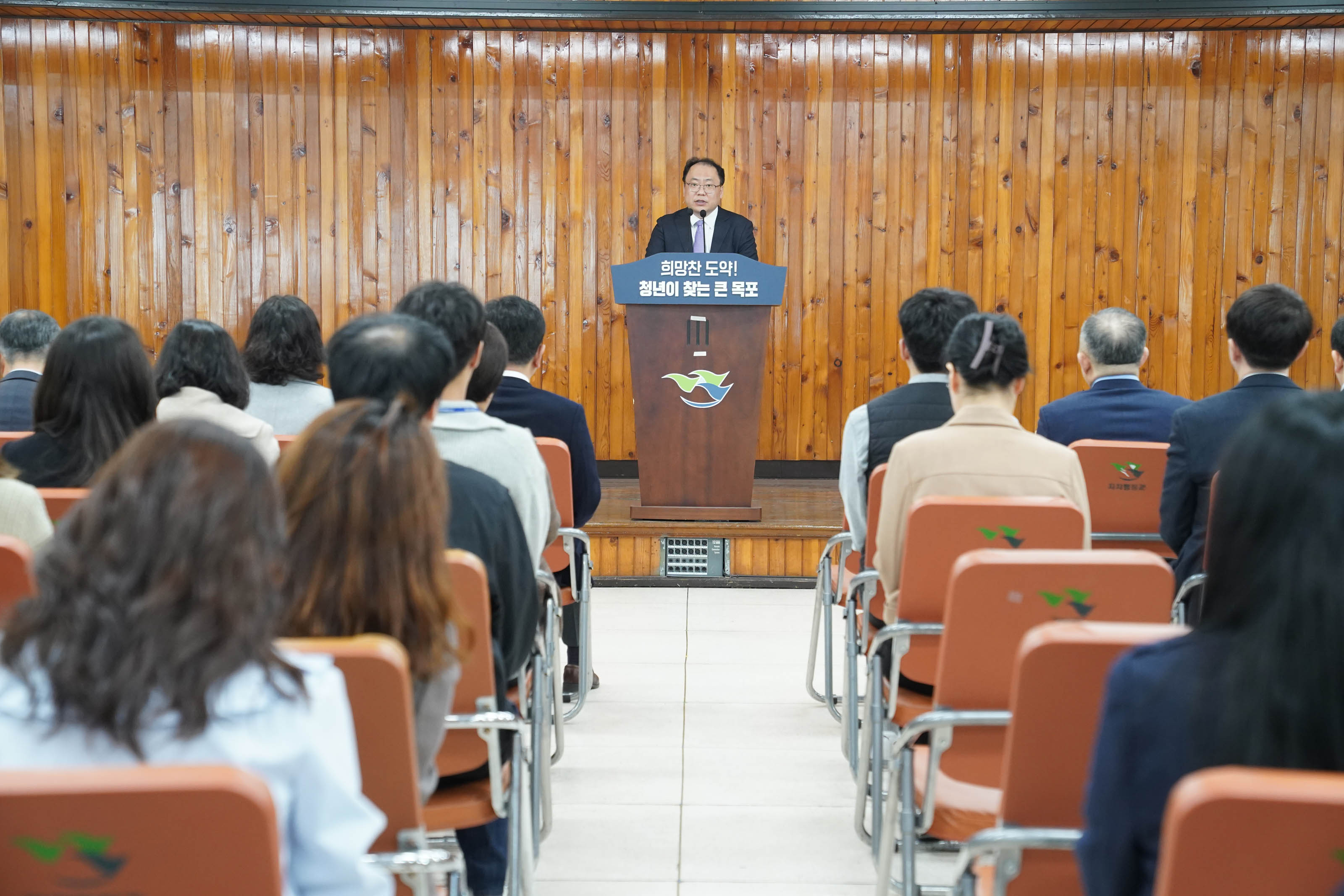 나무 패널 벽 앞 단상에서 정장을 입은 연설자가 발언 중이다. 단상에는 '희망찬 도약! 청년이 찾는 큰 목포'라는 문구가 쓰여 있고, 단상 아래에는 목포시 로고가 붙어 있다. 앞줄에는 청중들이 질서 있게 앉아 있으며, 의자 뒷면에는 '목포시' 문구와 로고가 인쇄되어 있다.