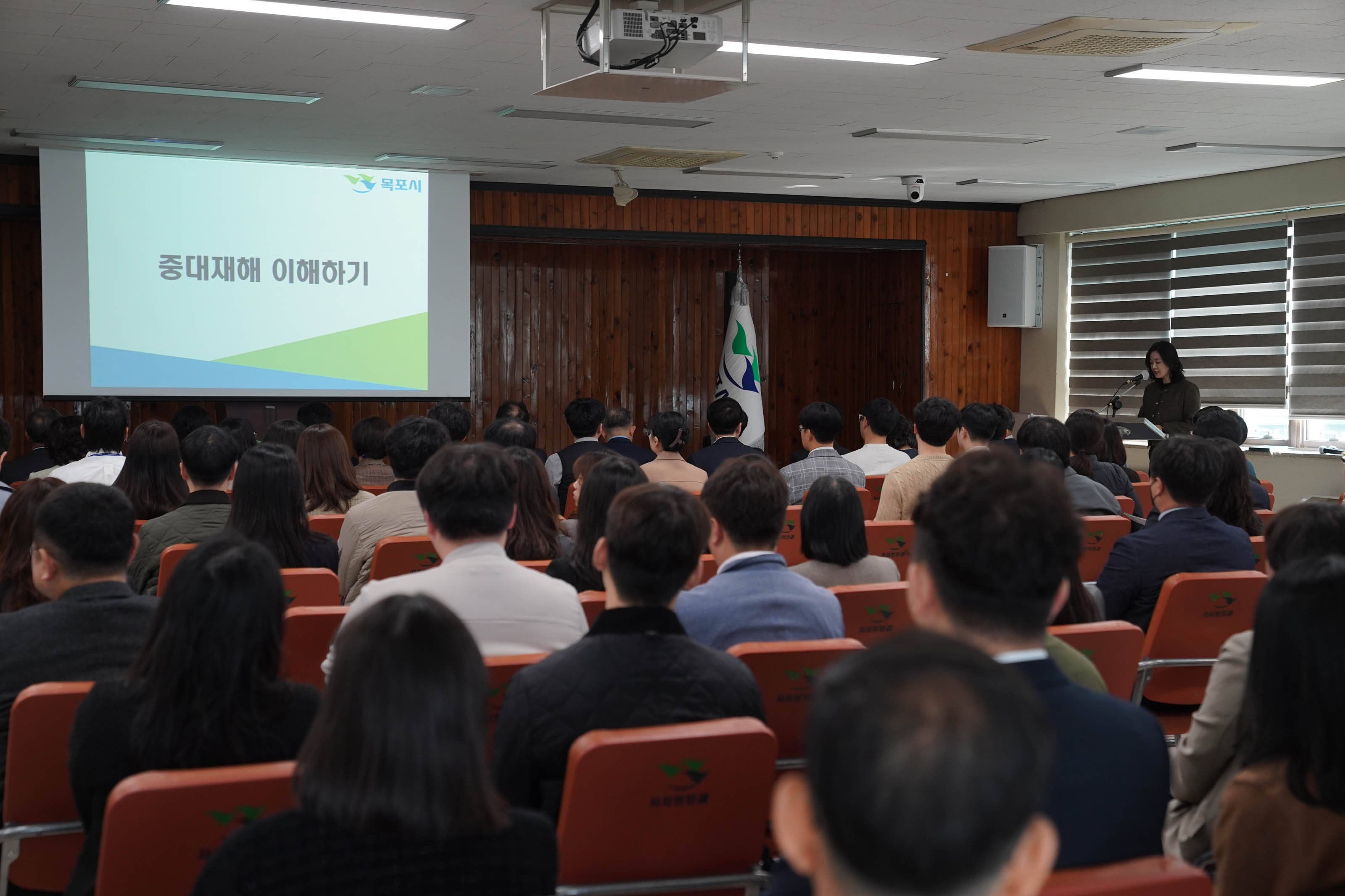 목포시에서 주관한 '중대재해 이해하기' 교육 현장. 스크린에는 '목포시' 로고와 함께 '중대재해 이해하기'라는 제목이 표시되어 있으며, 오른쪽 연단에서 강연자가 마이크를 통해 발표 중이다. 청중은 모두 정면을 바라보며 앉아 있고, 의자 뒷면에는 '목포시' 로고가 인쇄되어 있다. 강연장은 나무 패널 벽과 블라인드가 설치된 창문이 있는 구조이다.