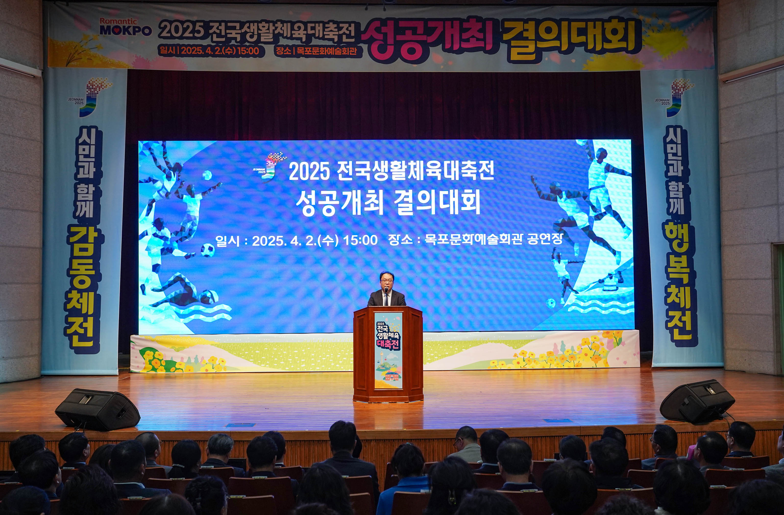 '2025전국생활체육대축전 성공개최 결의대회' 현수막이 위쪽에 설치되어 있고 '시민과 함께 감동체전', '시민과 함께 행복체전' 현수막이 스크린 양쪽으로 설치된 강당에서 '2025 전국생활체육대축전
성공개최 결의대회 일시:2025.4.2.(수) 15:00 장소: 목포문화예술회관 공연장' 문구가 적힌 스크린 앞에서 발표하는 관계자 그리고 지켜보는 사람들의 모습