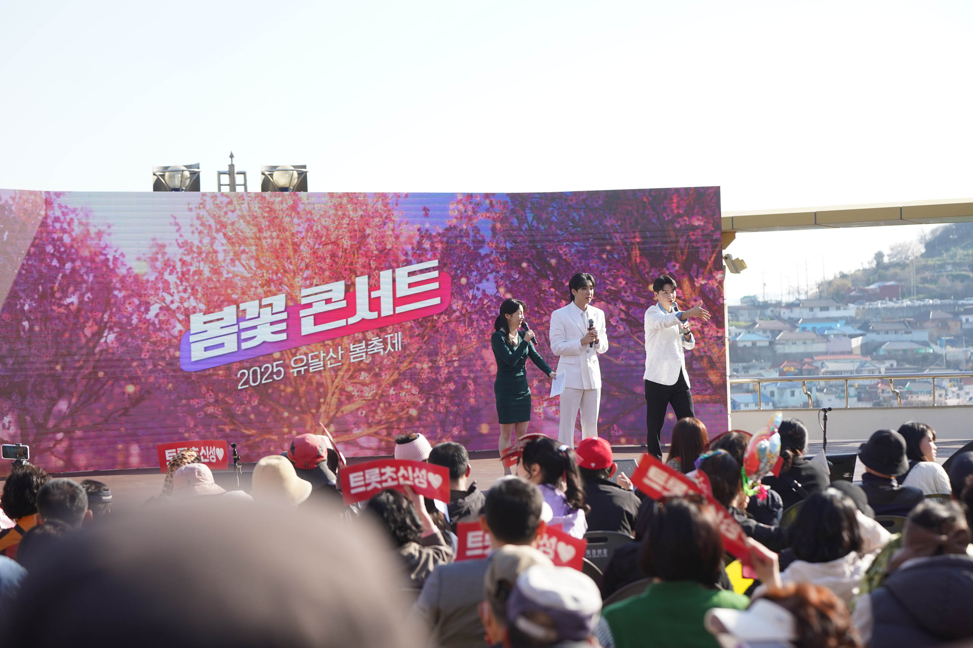 '봄꽃 콘서트 2025 유달산 봄축제' 문구가 띄워진 스크린 앞에서 노래하는 가수들과 '트롯초선생' 이라는 문구가 적힌 팻말을 들고 구경하는 사람들
