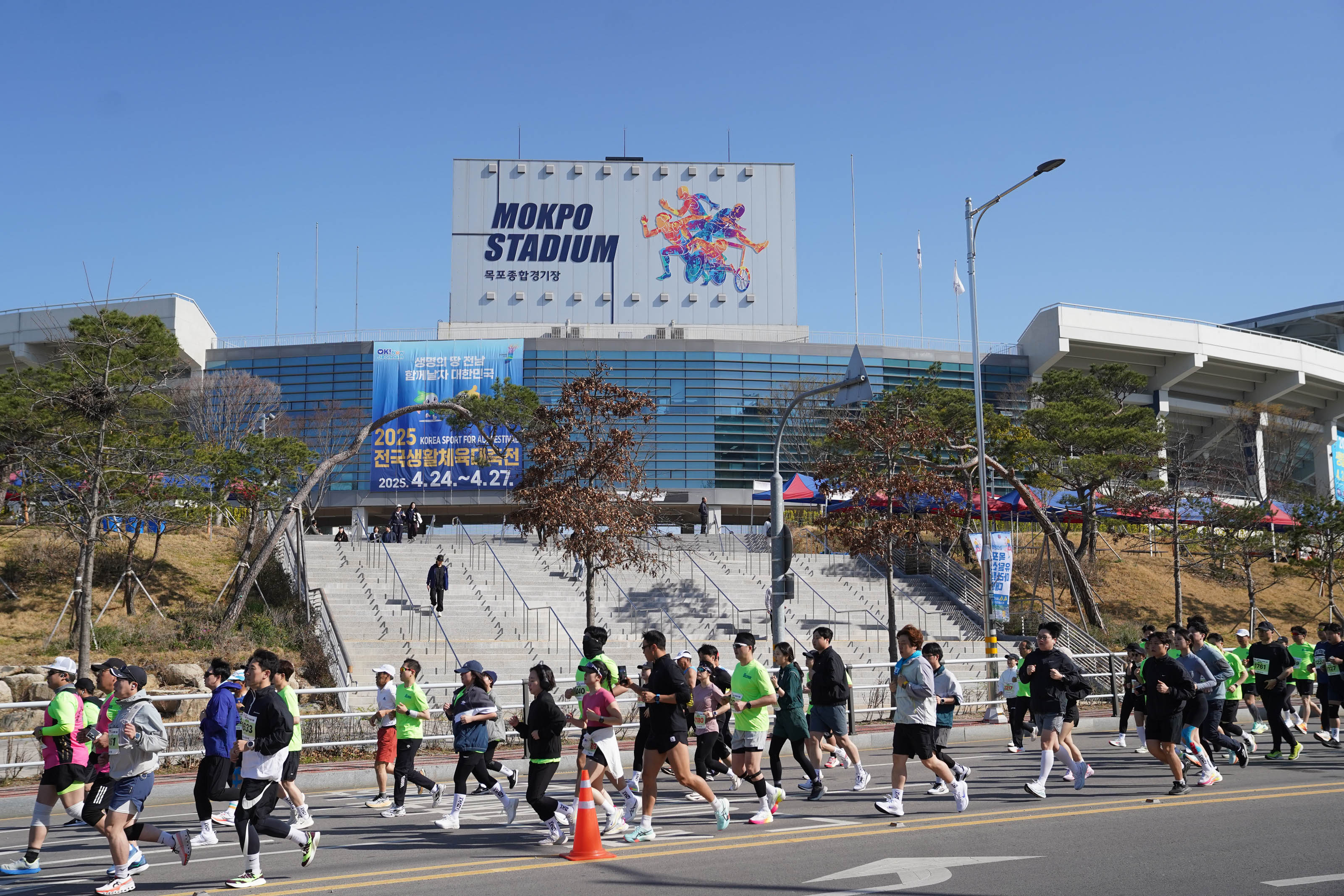 아스팔트 도로 위를 뛰어가는 참가자들의 모습. 배경으로 '2025 전국생활체육대축전 2025.4.24 ~ 4.27.' 문구가 적힌 현수막이 걸려있는 MOKPO STADIUM 목포종합경기장이 보임