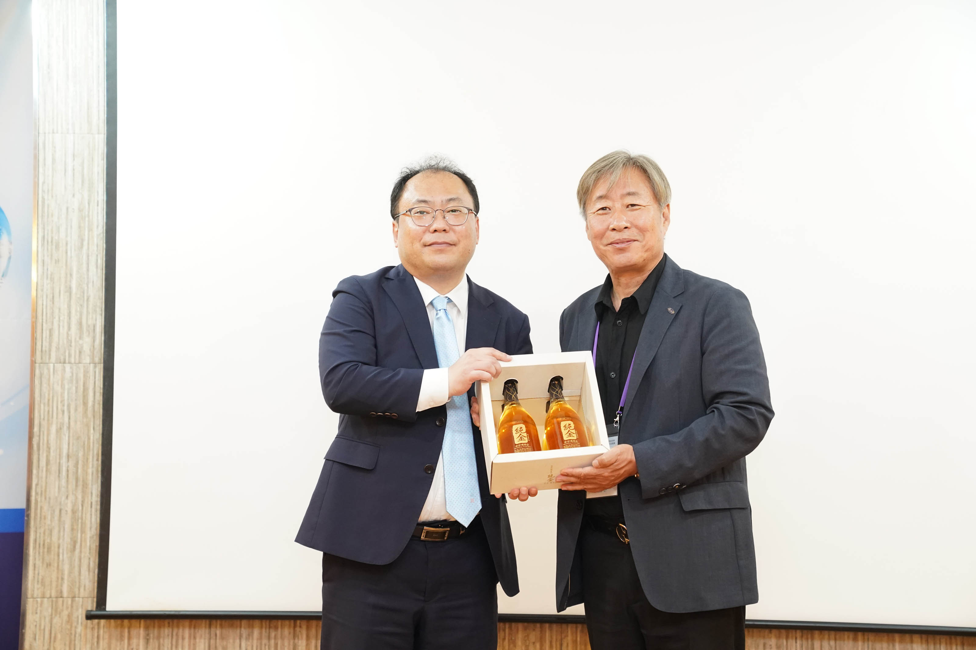 두 남성이 나란히 서서 두 병의 술이 들어있는 선물 세트를 함께 들고 기념 촬영을 하는 모습.