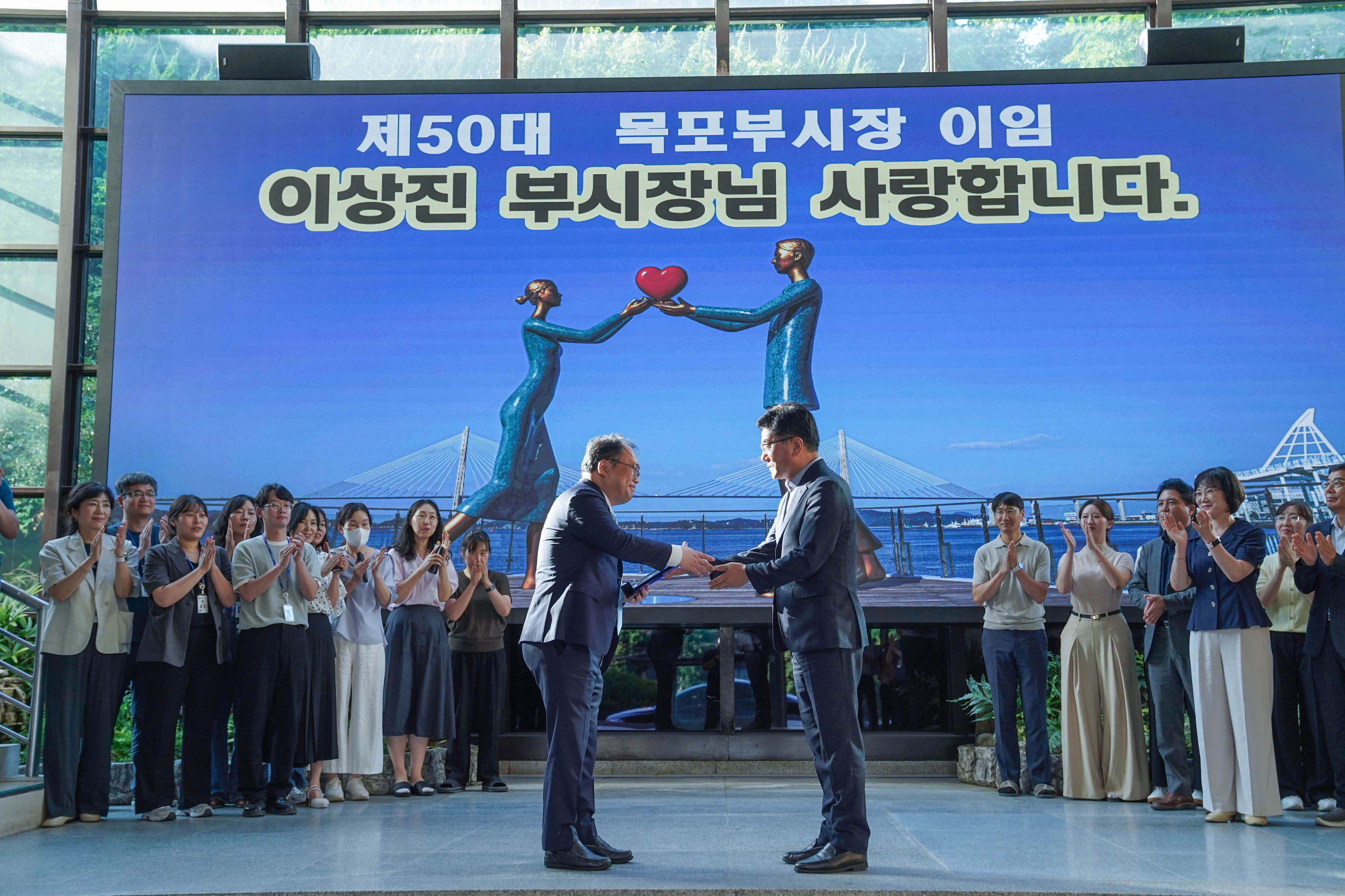대형 스크린에 '제50대 목포부시장 이임 이상진 부시장님 사랑합니다'라는 문구가 띄워진 가운데, 두 남성이 무대 중앙에서 기념품을 전달하며 악수하는 모습.