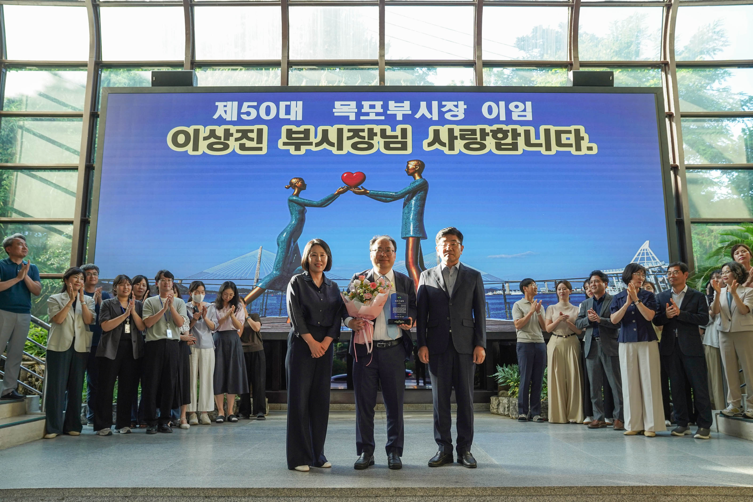 '제50대 목포부시장 이임 이상진 부시장님 사랑합니다' 문구가 있는 스크린 앞에서 이상진 부시장과 관계자들이 꽃다발과 기념패를 들고 정면을 바라보며 기념 촬영을 하는 모습.