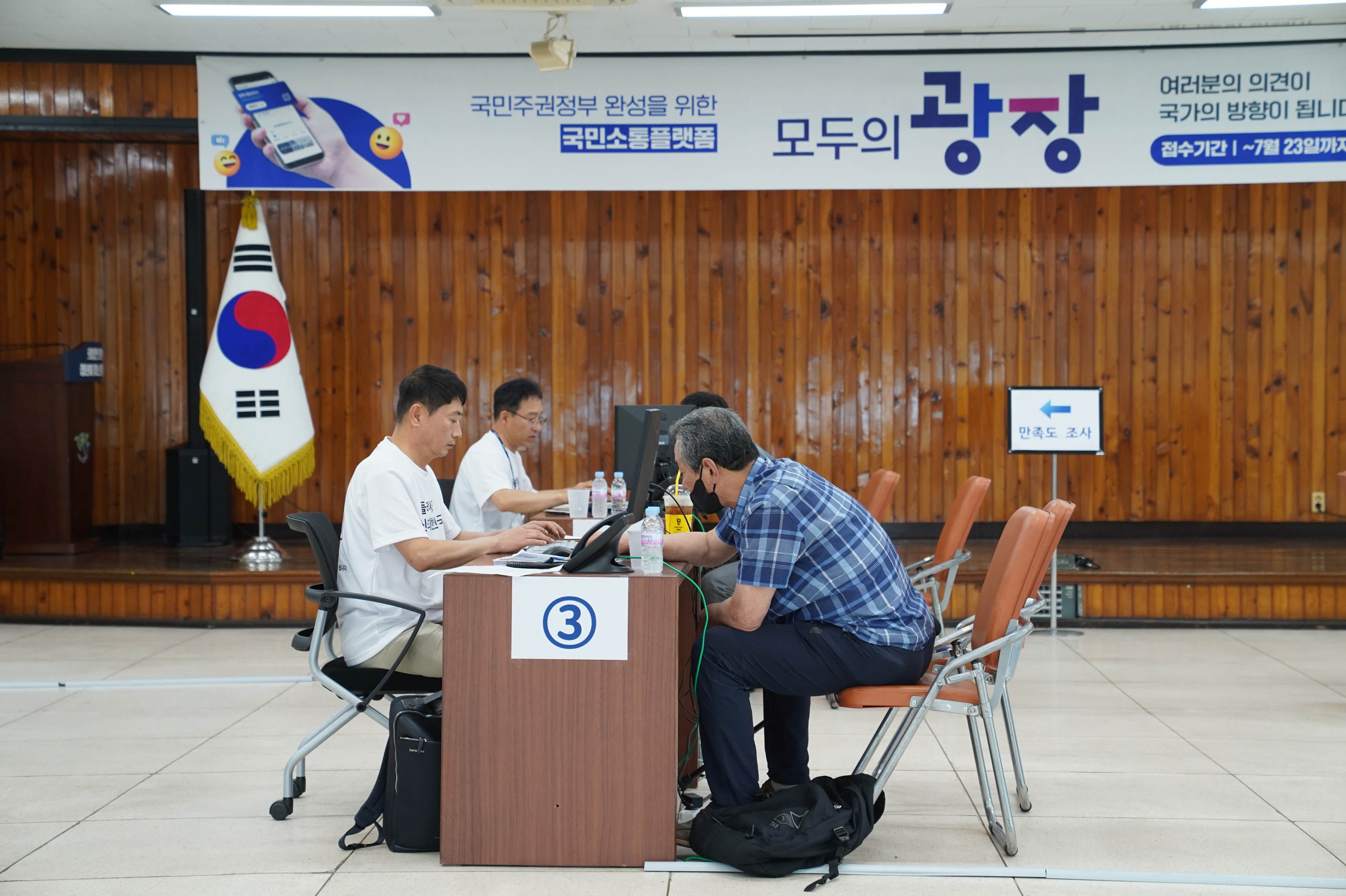 실내 행사장에서 '국민주권정부 완성을 위한 국민소통플랫폼 모두의 광장' 현수막 아래, 3번 테이블에 앉은 스태프가 민원인과 상담하며 컴퓨터를 조작하는 모습. 우측에는 '만족도 조사' 안내판이 있음.