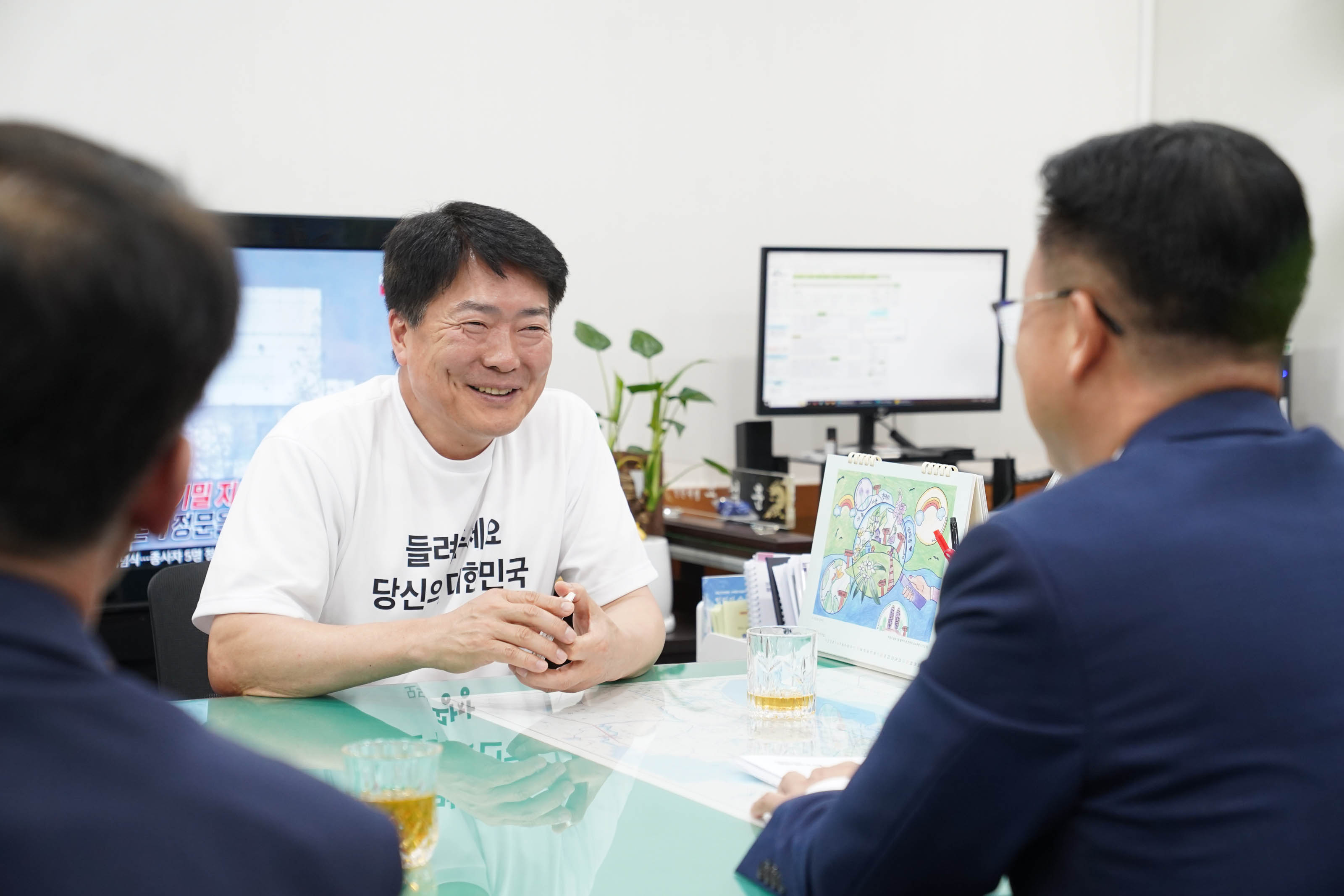 '들려주세요 당신의 대한민국' 문구가 적힌 흰색 티셔츠를 입은 남성이 회의 테이블에 앉아 밝게 웃으며 맞은편 사람과 대화하는 모습. 테이블 위에는 그림이 그려진 탁상 달력과 찻잔이 놓여 있음.