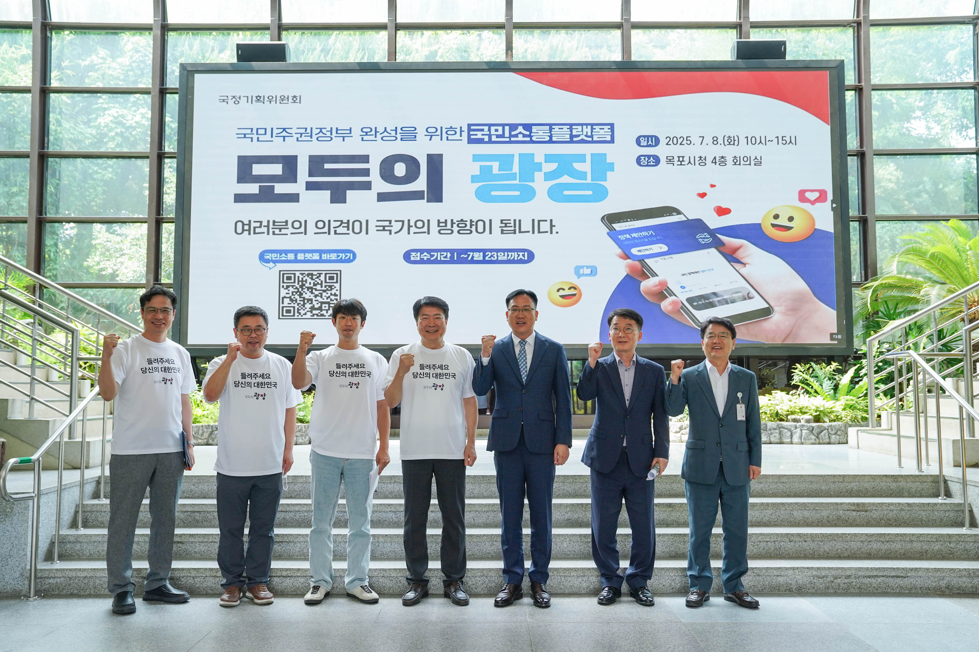 '국정기획위원회'의 '국민주권정부 완성을 위한 국민소통플랫폼 모두의 광장' 홍보 영상이 나오는 대형 스크린 앞에서 7명의 남성이 나란히 서서 주먹을 쥐고 파이팅 포즈를 취하고 있는 단체 사진.