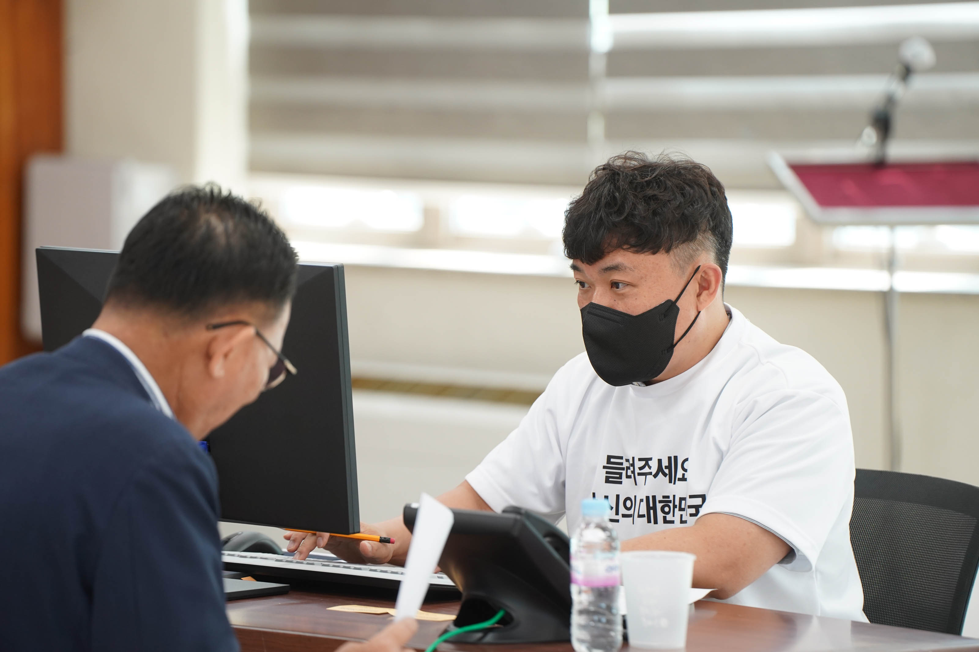 '들려주세요 당신의 대한민국' 문구가 적힌 흰색 티셔츠를 입은 스태프가 검은색 마스크를 쓰고 모니터 앞에서 민원인과 대화에 집중하고 있는 상반신 모습.