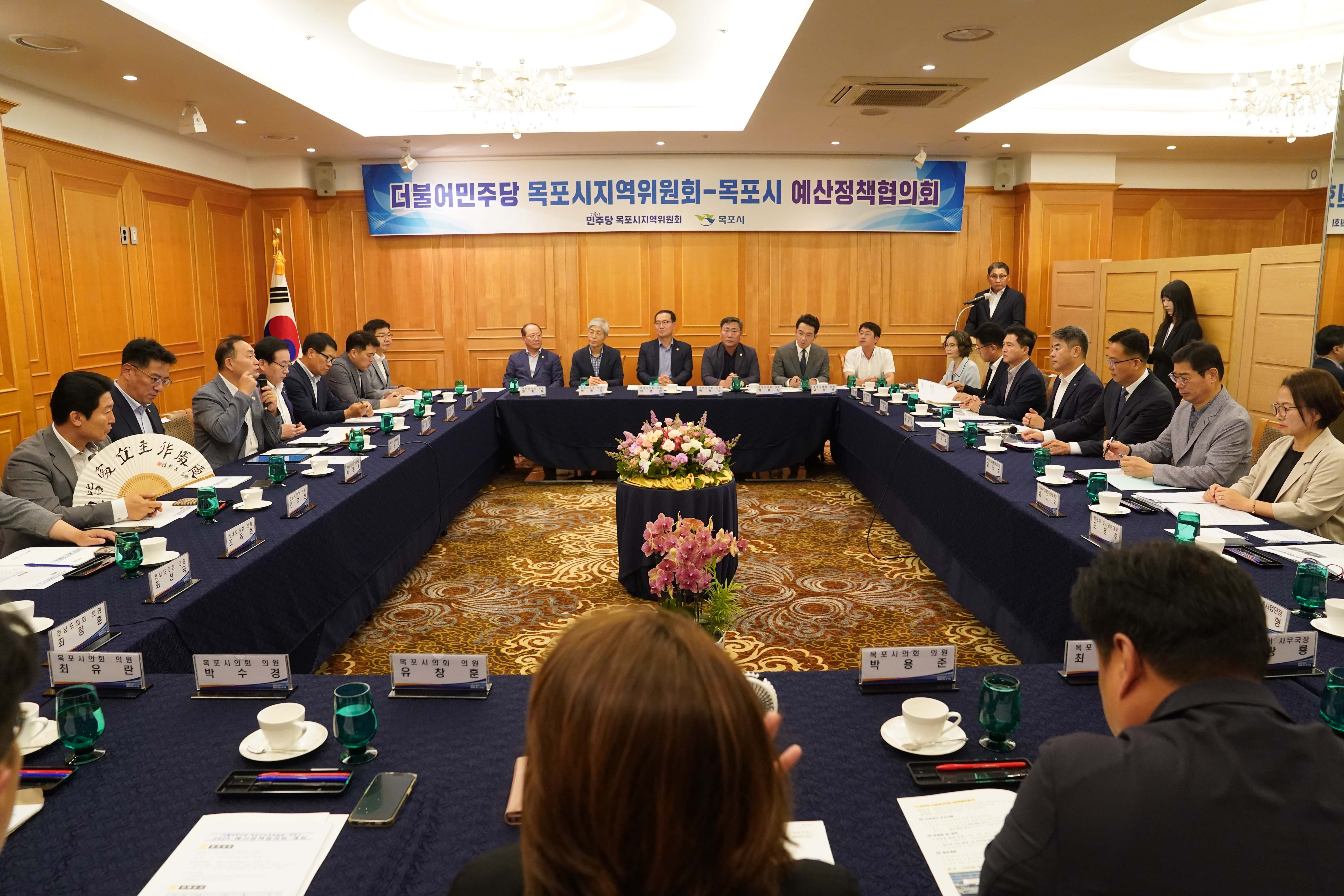 '더불어민주당 목포시지역위원회-목포시 예산정책협의회' 현수막이 걸린 회의장 전경. 정장을 입은 관계자들이 대형 테이블에 둘러앉아 있으며, 테이블 위에는 꽃바구니와 찻잔, 회의 자료가 배치되어 있습니다.