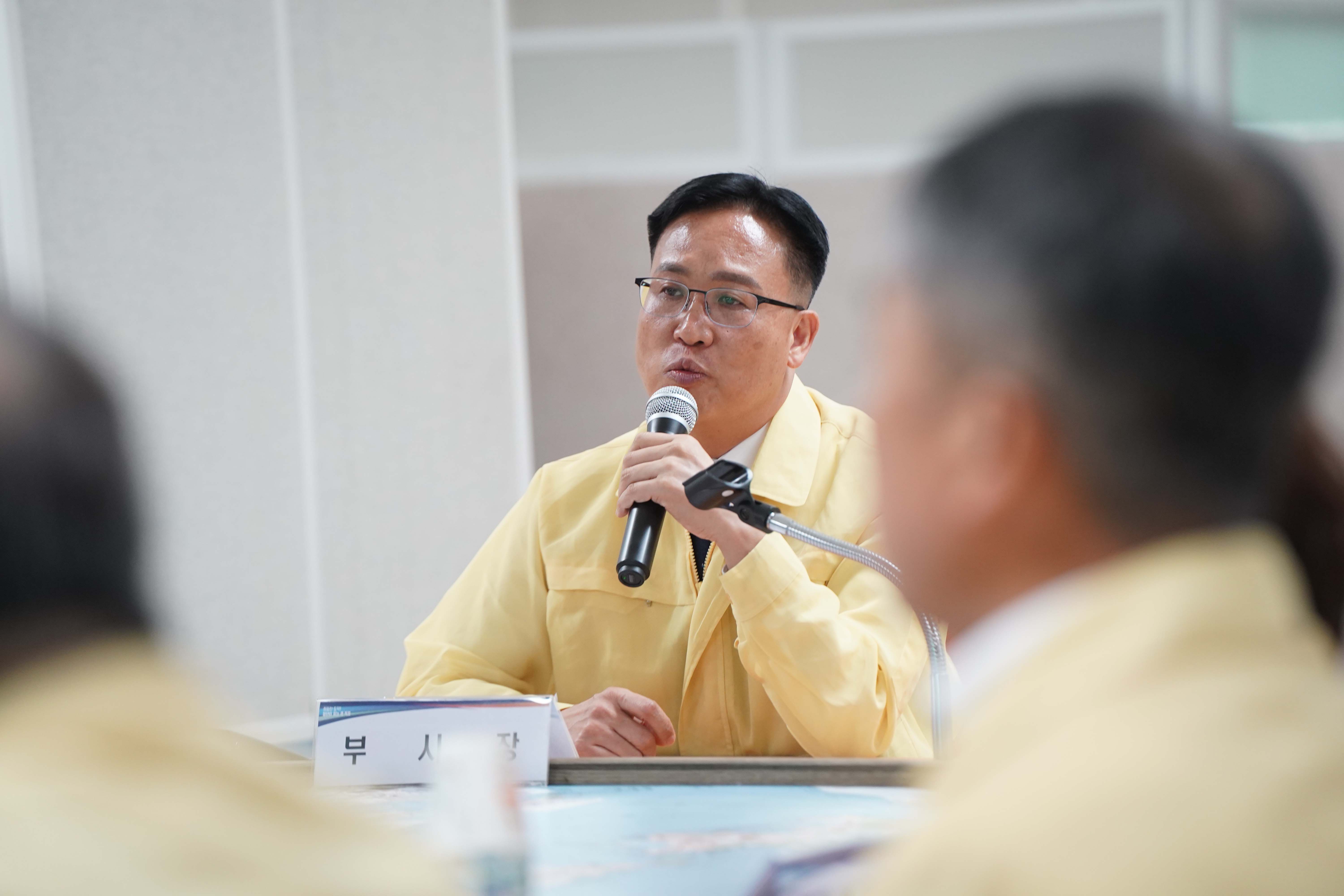 '부시장'이라는 명패가 놓인 책상 앞에서 안경을 쓰고 민방위복을 입은 남성이 마이크를 잡고 발언하는 근접 촬영 사진.