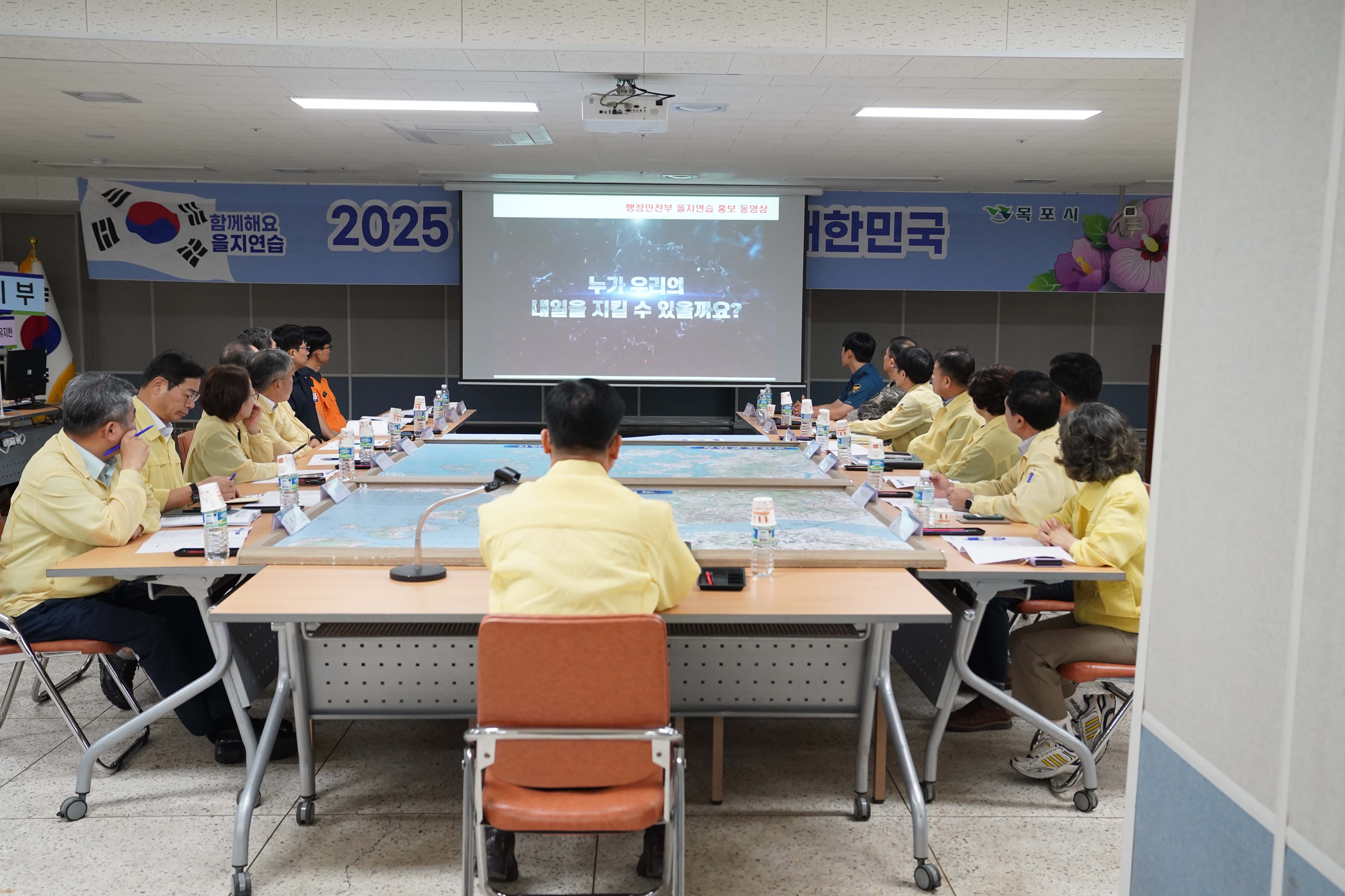 대형 지도가 펼쳐진 회의 탁자에 공무원들이 둘러앉아 정면의 스크린을 시청하고 있는 뒷모습. 스크린에는 '행정안전부 을지연습 홍보 동영상'이 재생 중이며, 왼쪽 벽면에는 태극기와 함께 '함께해요 을지연습' 현수막이 붙어 있음.