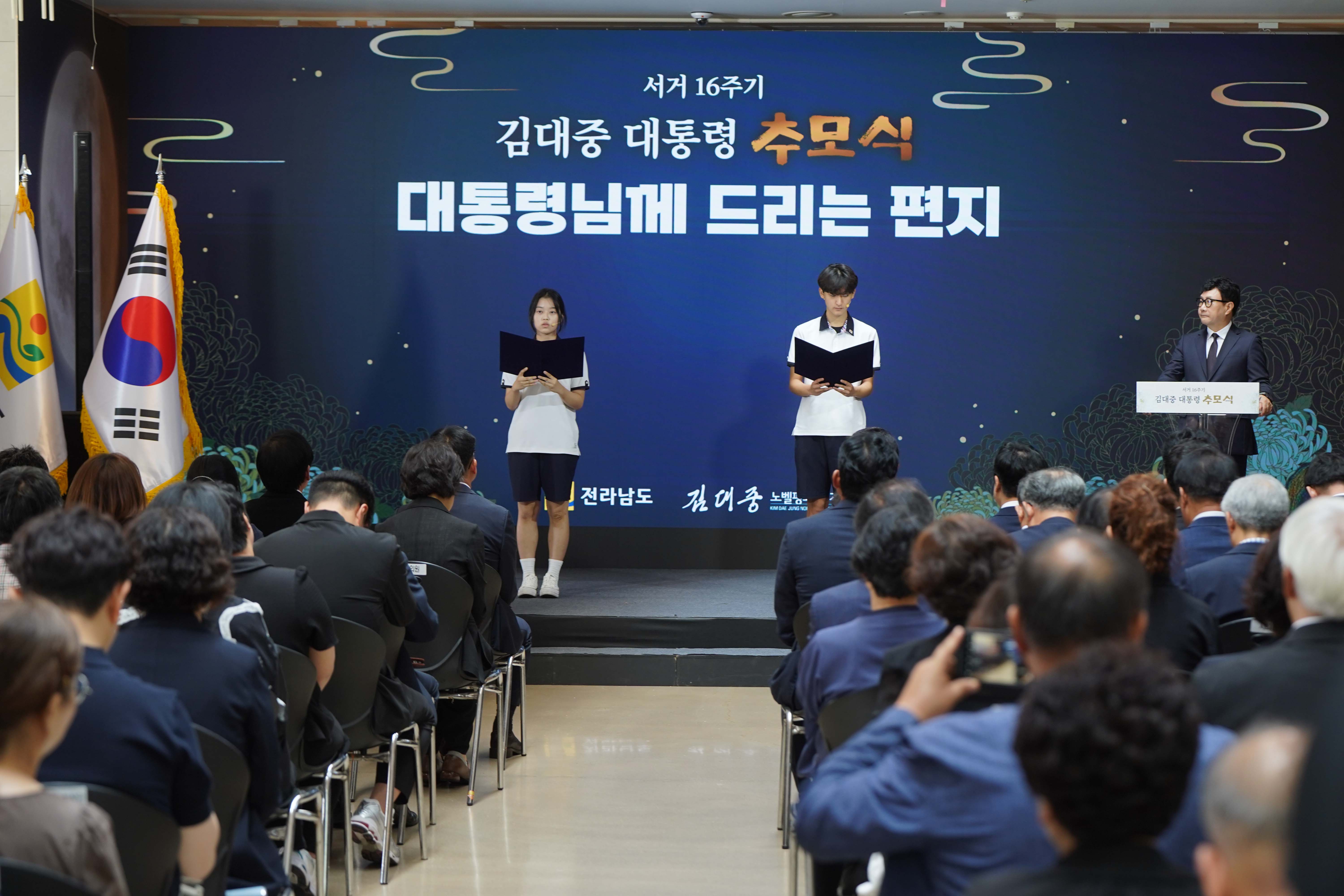 추모식 프로그램 중 '대통령님께 드리는 편지' 순서. 두 명의 학생이 무대 위에서 편지를 낭독하고 있으며, 하단에 전라남도와 김대중 노벨평화상 기념관 로고가 보임.