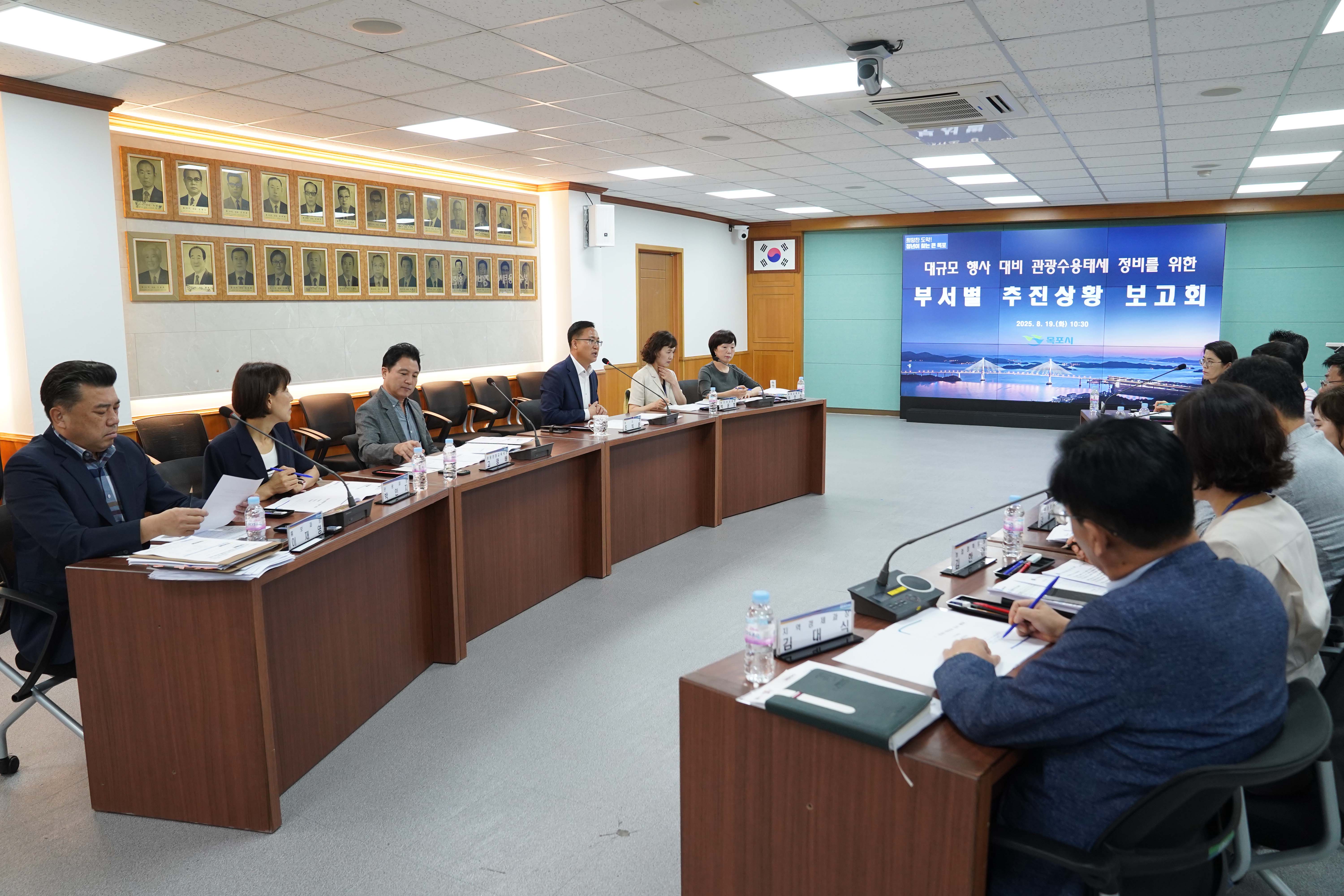 회의실에서 진행 중인 '부서별 추진상황 보고회' 모습. 왼쪽 벽면 상단에 역대 시장 및 군수들의 인물 사진 액자가 줄지어 걸려 있고, 오른쪽 스크린에는 '대규모 행사 대비 관광수용태세 정비를 위한 부서별 추진상황 보고회', '2025. 8. 19.(화) 10:30', 목포시 로고가 보임. 테이블 위 명패에는 '주택과장 김대식' 등의 텍스트가 식별됨.