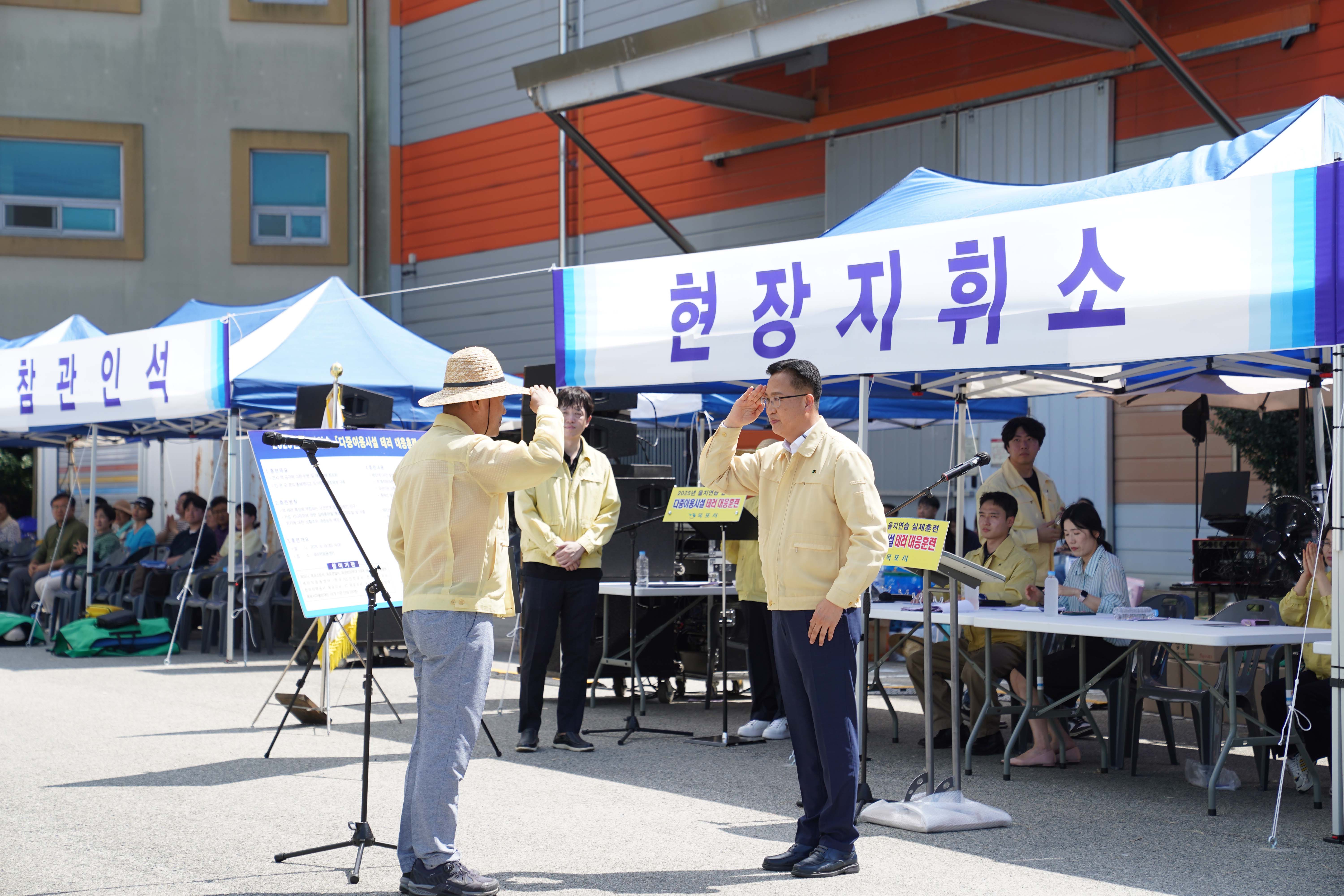 '현장지휘소' 천막 앞에서 훈련 참가자가 지휘관에게 거수경례로 보고하는 모습. 배경에 '다중이용시설 테러 대응훈련' 안내판이 있음.
