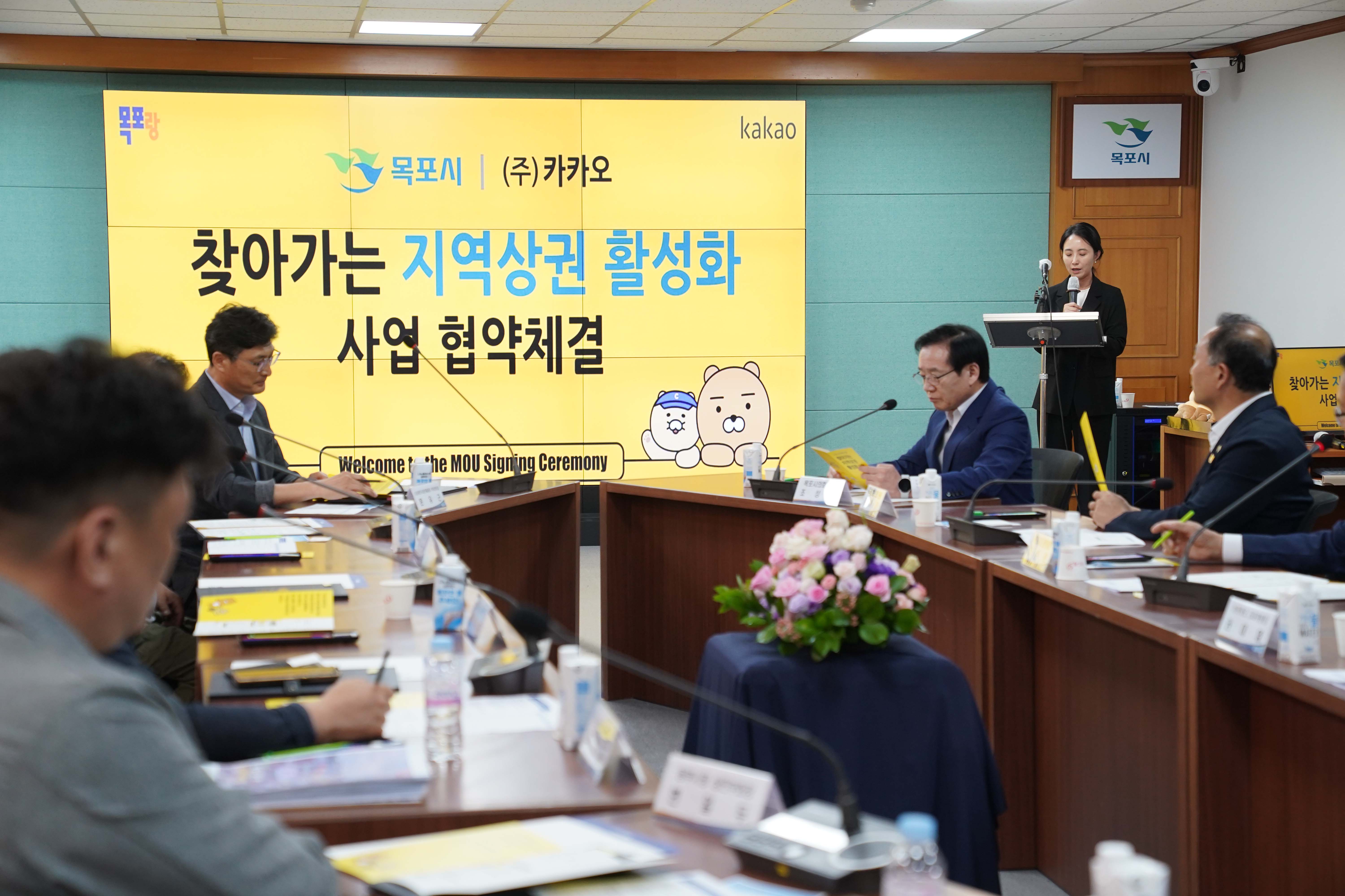 회의실 단상에서 한 여성이 사회를 보고 있으며, 중앙 스크린에는 '목포시 | (주)카카오 찾아가는 지역상권 활성화 사업 협약체결' 문구가 크게 노출됨. 테이블 위에는 꽃바구니가 놓여 있음.
