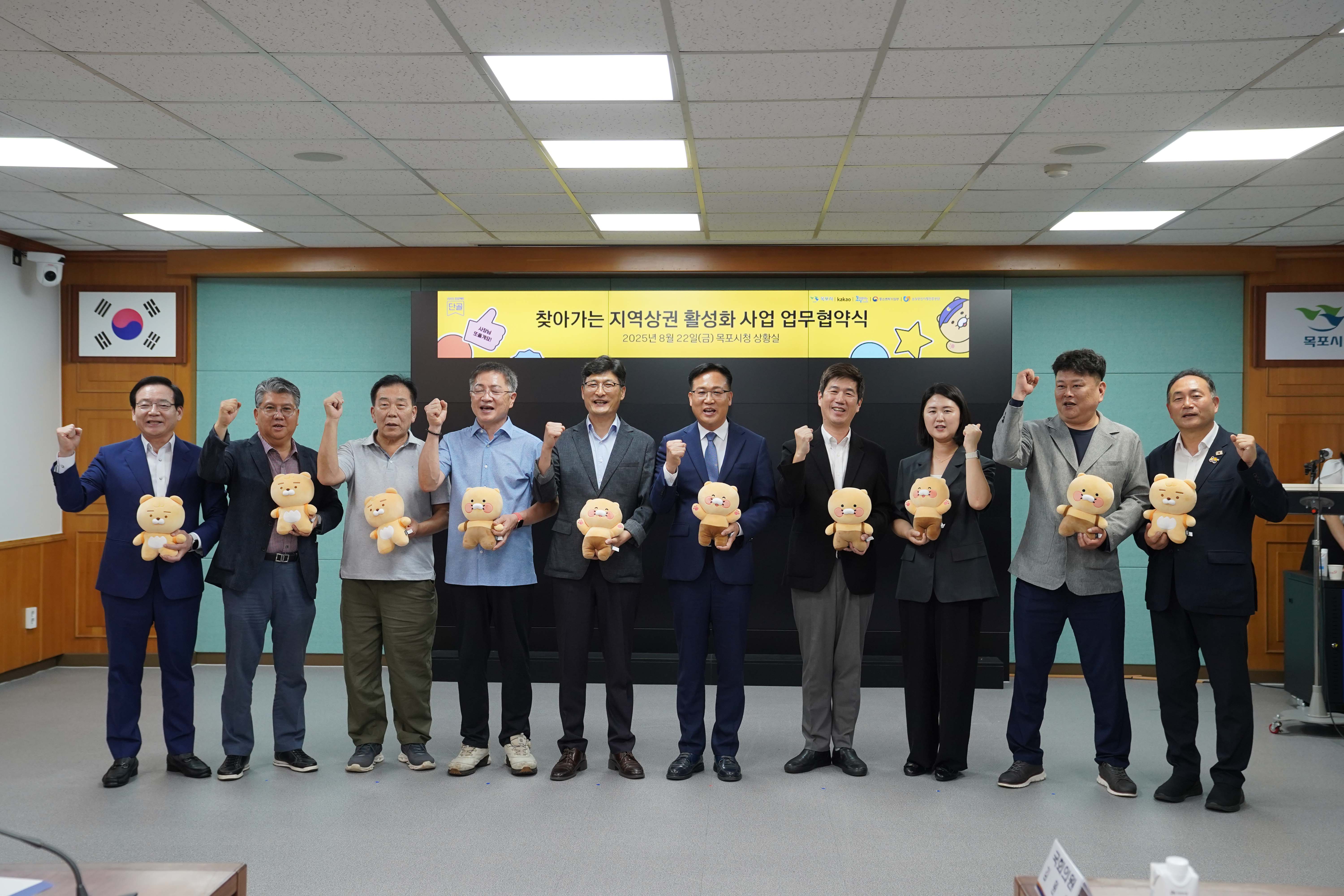 업무협약식 기념 촬영 중, 참석자들이 한 손에는 카카오프렌즈 라이언 인형을 들고 다른 한 손은 파이팅 포즈를 취하며 웃고 있는 모습. 상단에는 '찾아가는 지역상권 활성화 사업 업무협약식' 문구가 보임.
