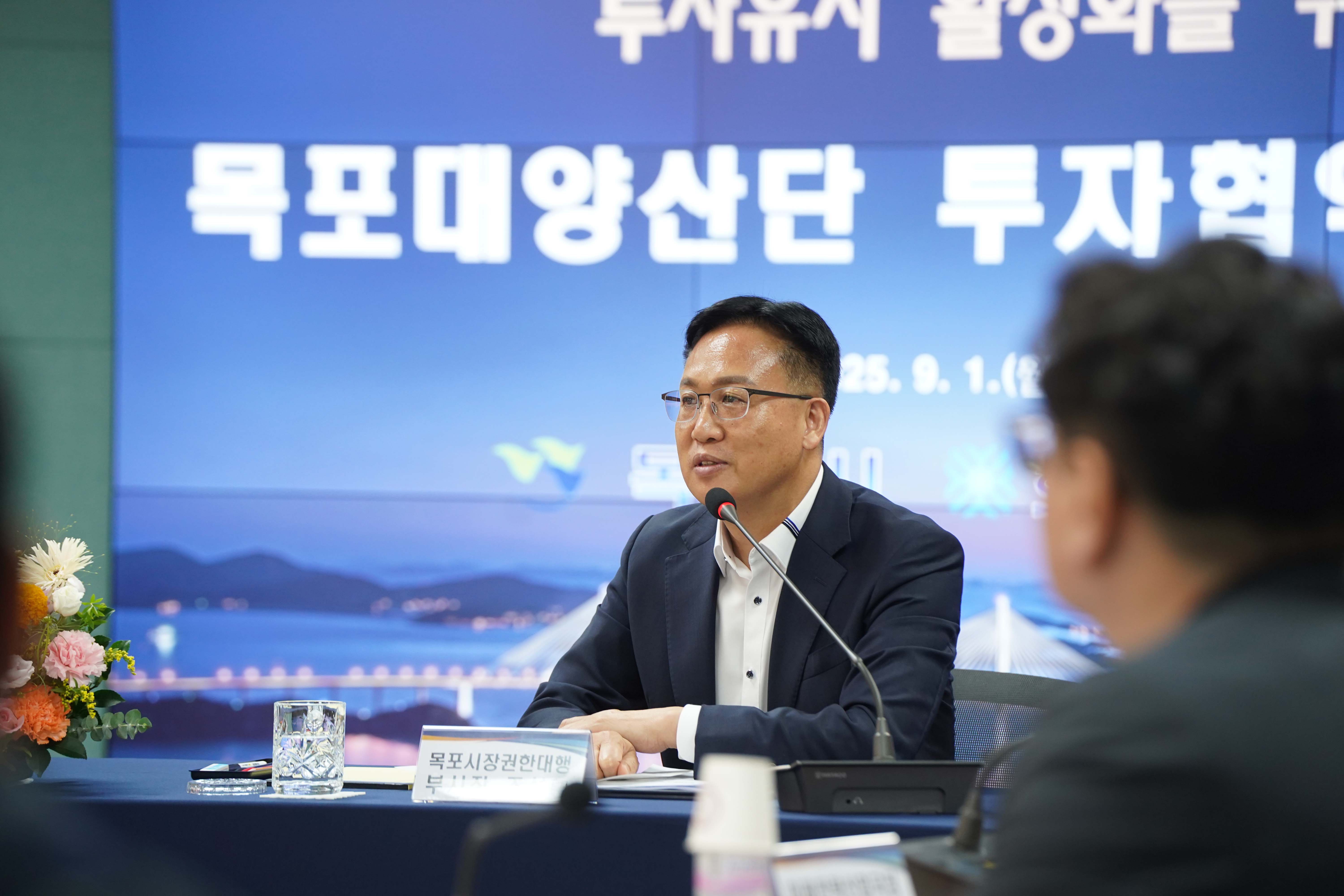 '목포시장권한대행 부시장 조석훈' 명패 뒤에 앉은 조석훈 부시장이 안경을 쓰고 마이크 앞에서 발언하는 모습. 배경 스크린에는 '목포대양산단 투자협약' 문구가 일부 보임.