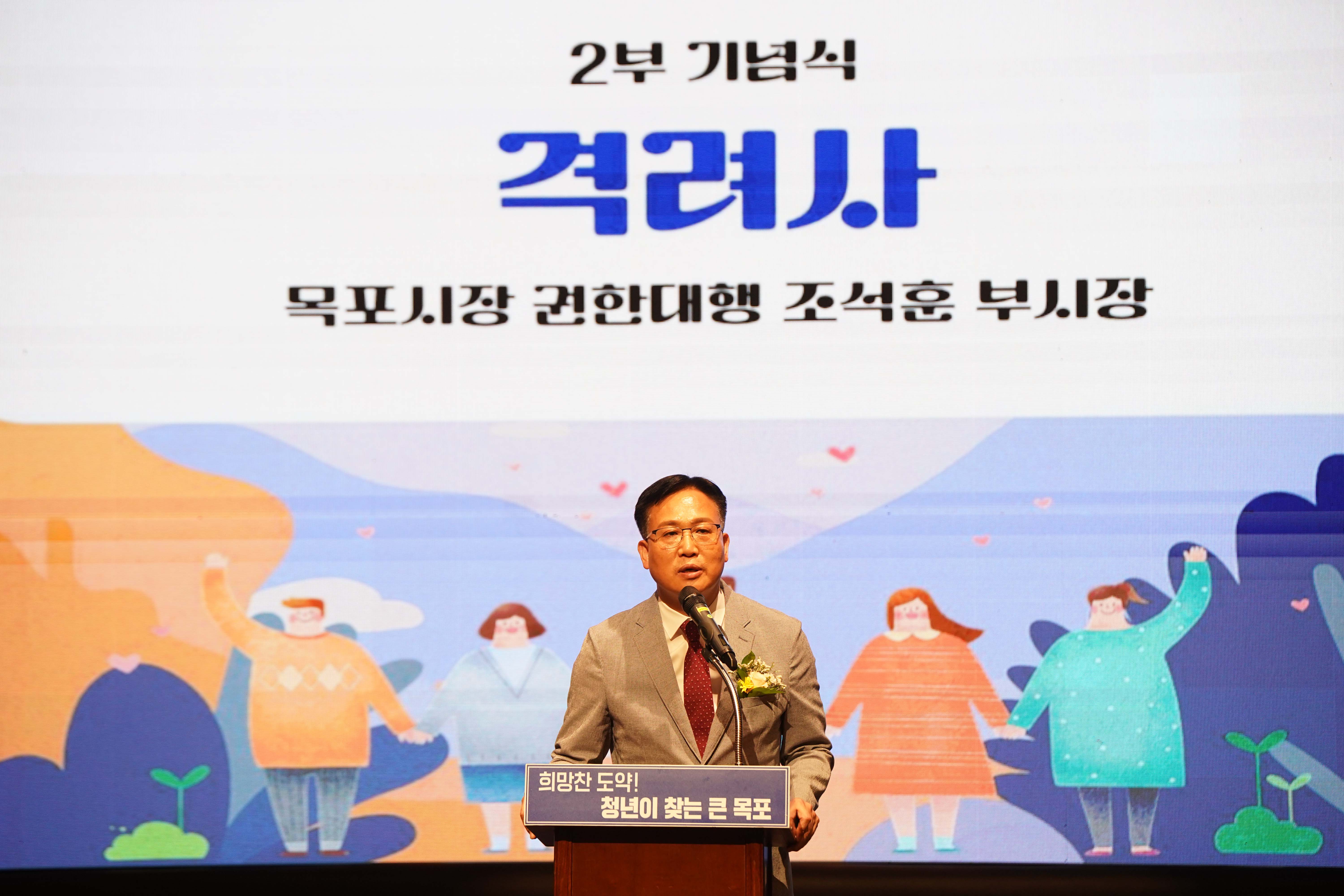 조석훈 부시장이 '희망찬 도약! 청년이 찾는 큰 목포'라고 적힌 단상 뒤에서 격려사를 하는 모습. 배경 스크린에는 '2부 기념식 격려사 목포시장 권한대행 조석훈 부시장' 문구와 함께 다정한 사람들의 일러스트가 그려져 있음.