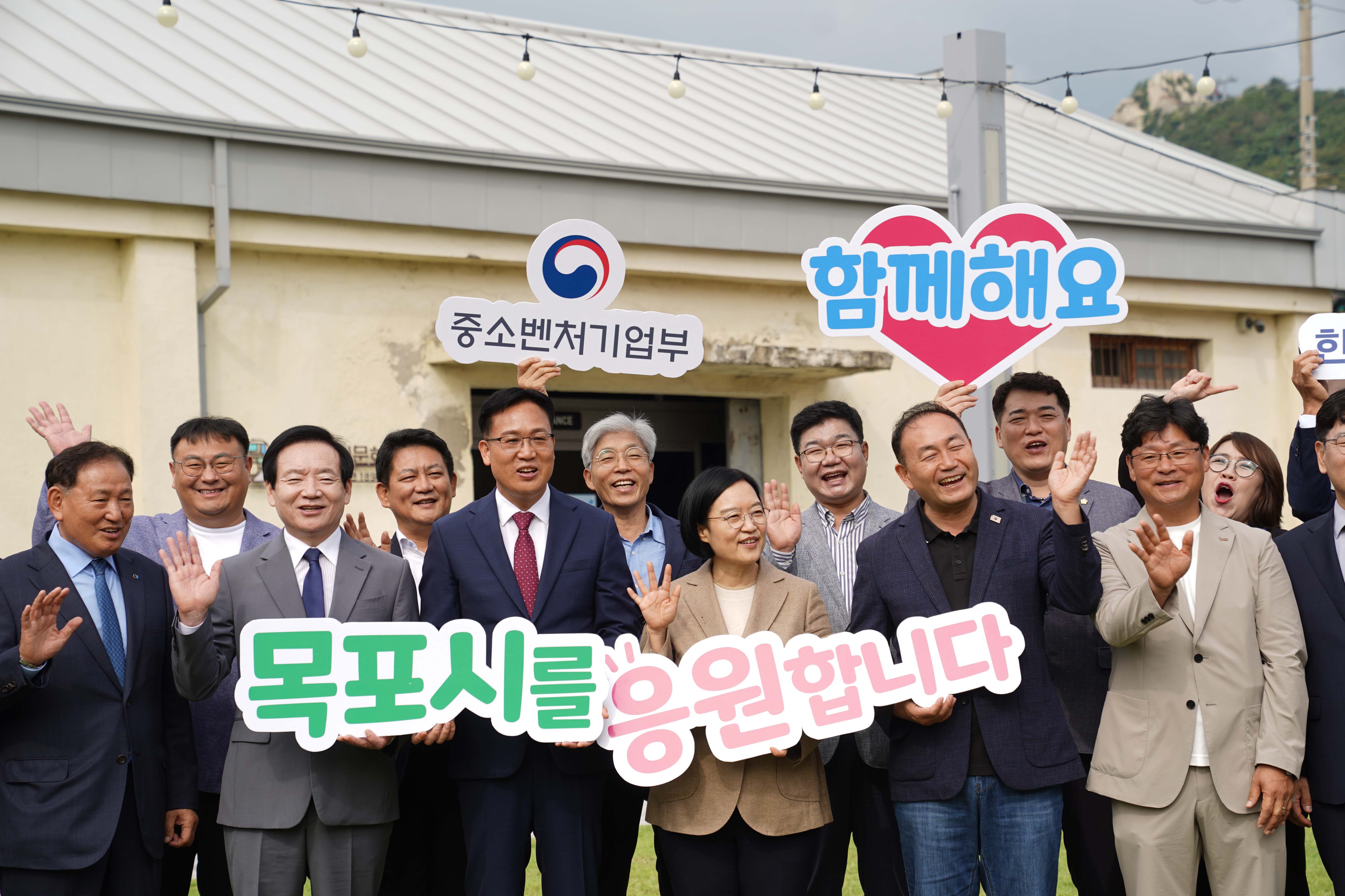 '중소벤처기업부', '함께해요', '목포시를 응원합니다'라는 문구의 피켓을 든 한성숙 장관과 관계자들이 환하게 웃으며 손을 흔드는 모습.
