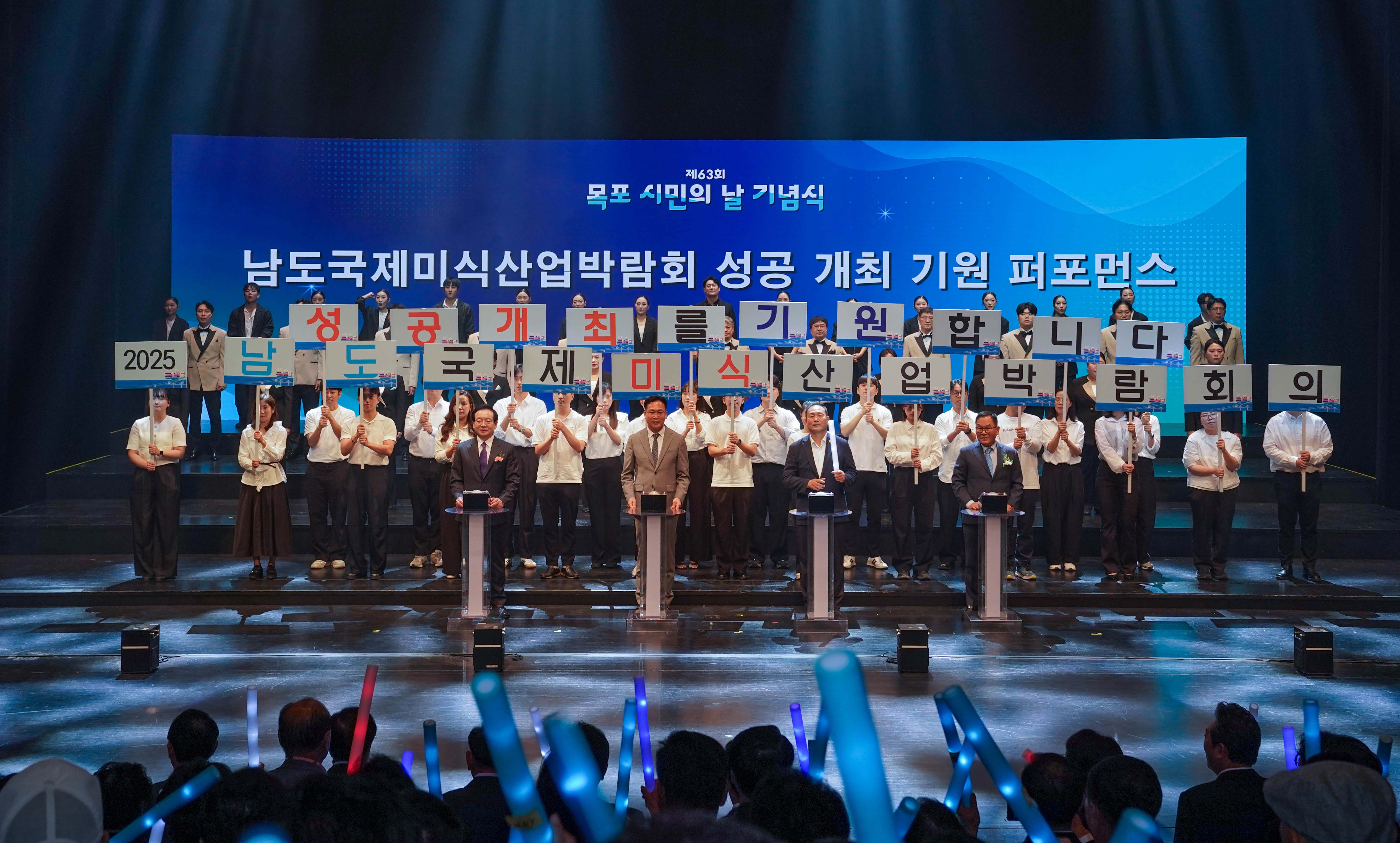 '제63회 목포 시민의 날 기념식' 무대에서 진행된 '남도국제미식산업박람회 성공 개최 기원 퍼포먼스'. 무대 위 사람들이 '성공 개최를 기원합니다', '2025 남도국제미식산업박람회' 문구가 적힌 글자 판넬을 들고 있으며, 앞줄 내빈들이 버튼 터치 퍼포먼스를 준비하고 있는 전경.
