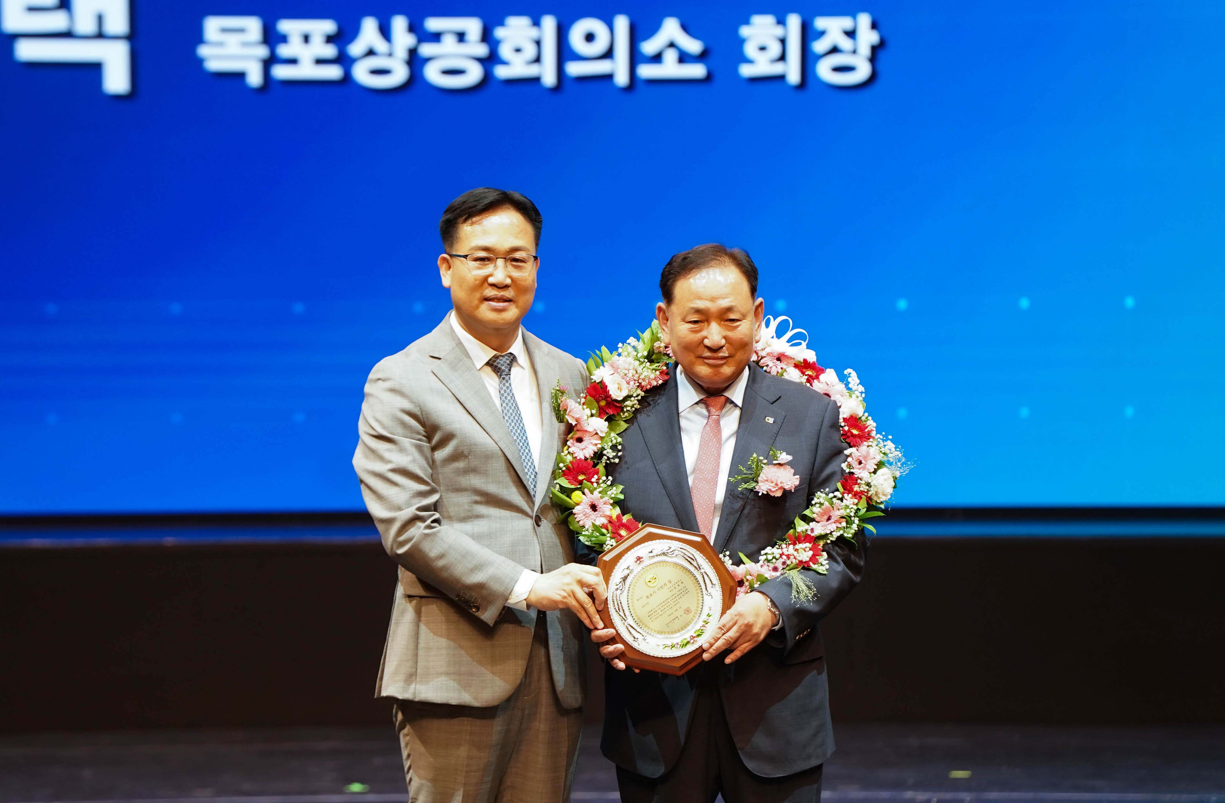 '시민의 상'을 수상한 정현택 목포상공회의소 회장과 조석훈 부시장이 함께 상패를 들고 기념 촬영을 하는 모습. 정현택 회장은 화려한 꽃목걸이를 걸고 있음.