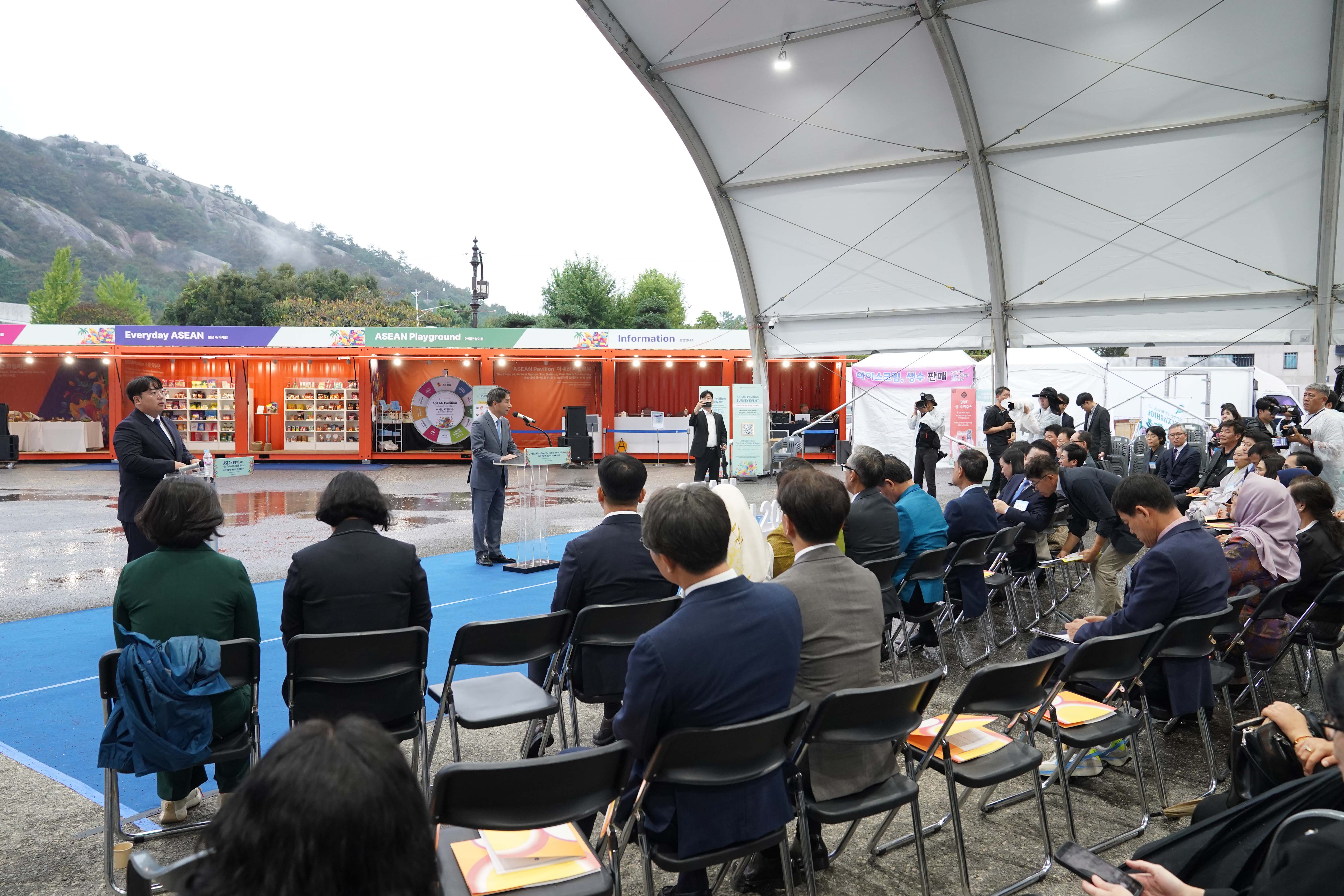 야외 행사 현장. 'Everyday ASEAN', 'ASEAN Playground', 'Information'이라고 적힌 주황색 컨테이너 부스 앞에서 한 남성이 연단에 서서 발언 중이며 관객들이 경청하고 있음.