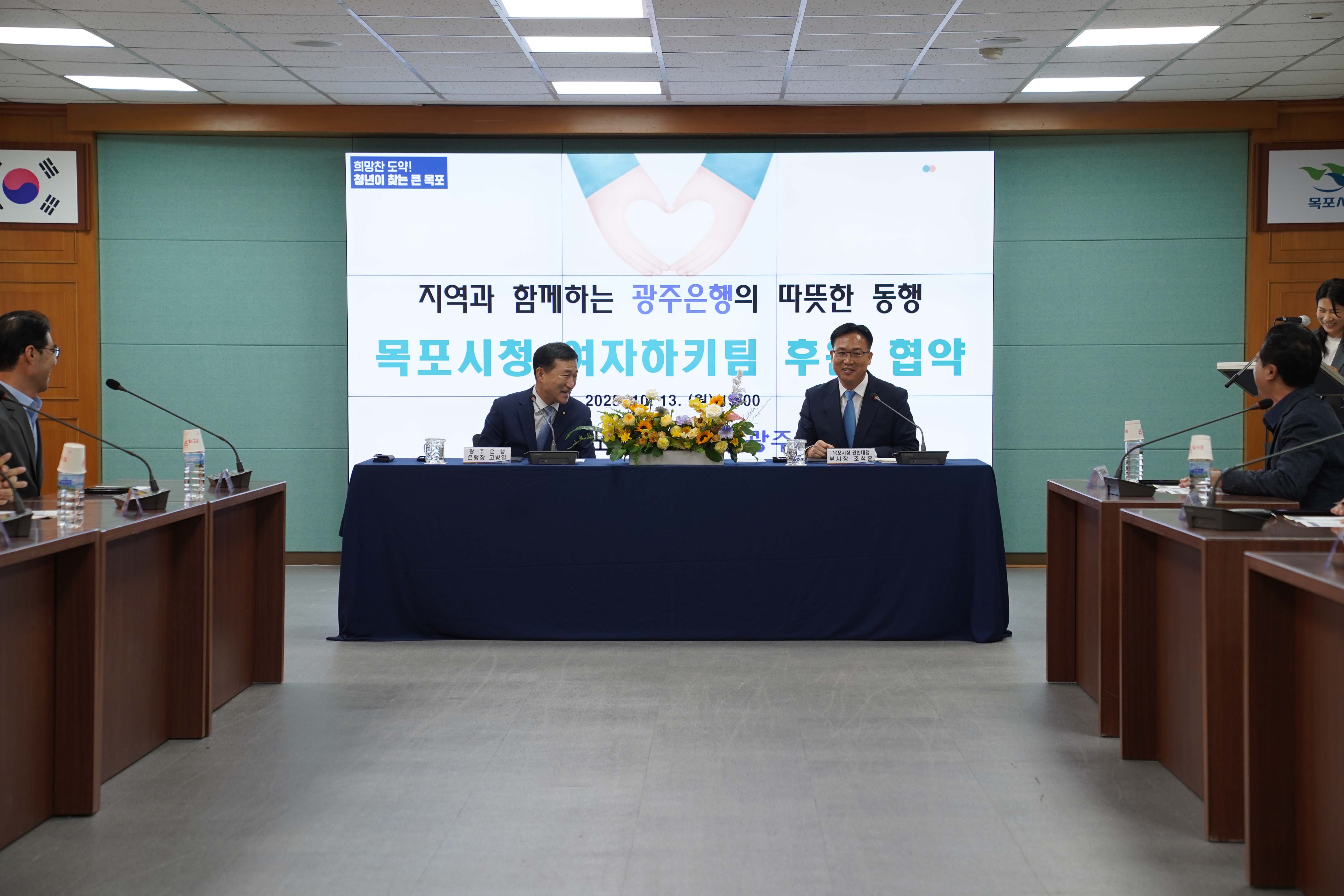 '지역과 함께하는 광주은행의 따뜻한 동행, 목포시청 여자하키팀 후원 협약' 행사장 전경. 중앙 테이블에 고병일 광주은행장과 조석훈 목포부시장이 마주 앉아 대화하는 모습.