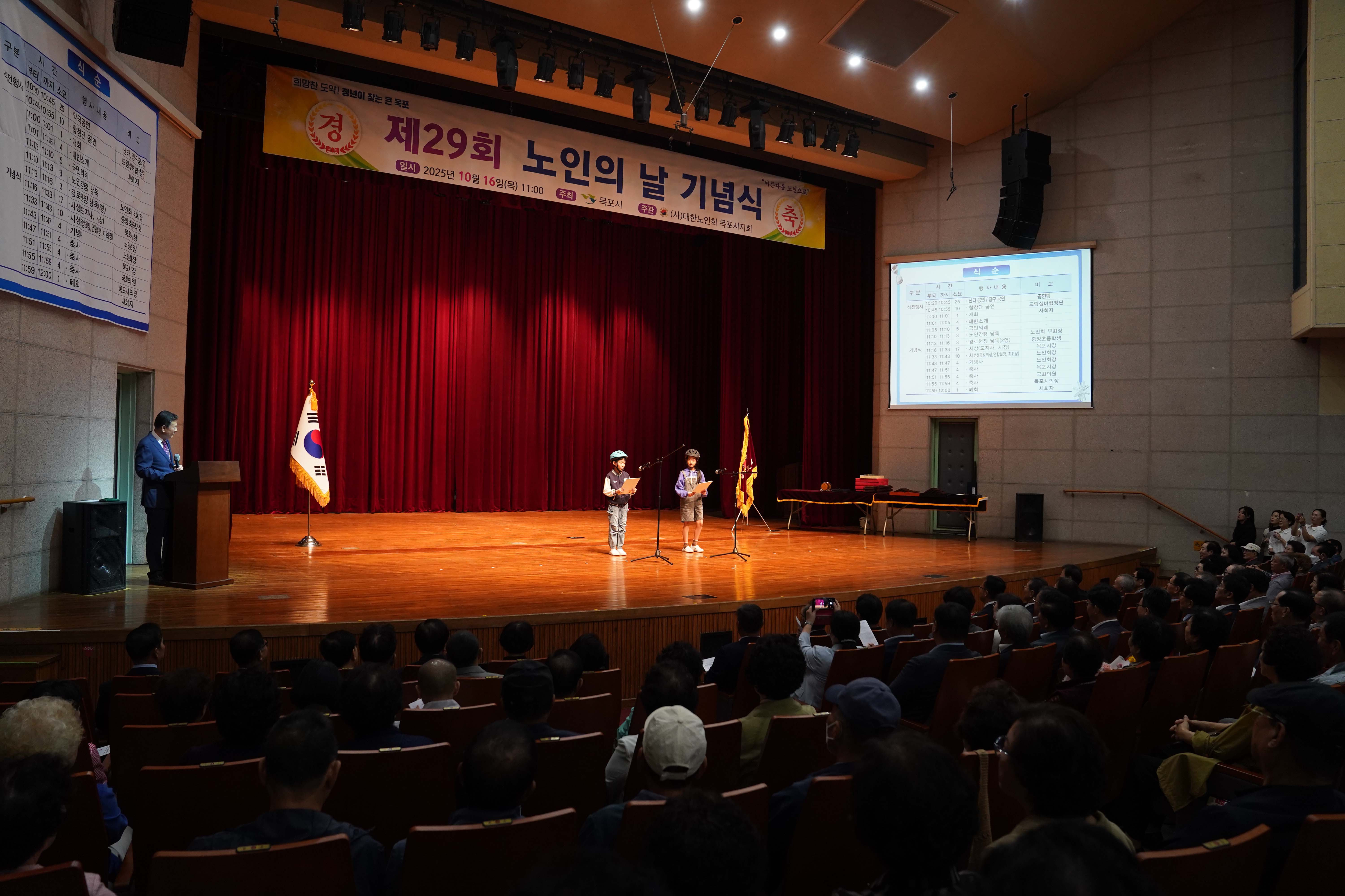 기념식 무대에서 헬멧을 쓴 두 어린이가 마이크 앞에 서서 발표를 하고 있는 모습. 우측 스크린에는 행사의 '식순'이 띄워져 있고, 좌석을 가득 채운 관람객들이 이를 지켜보고 있음.