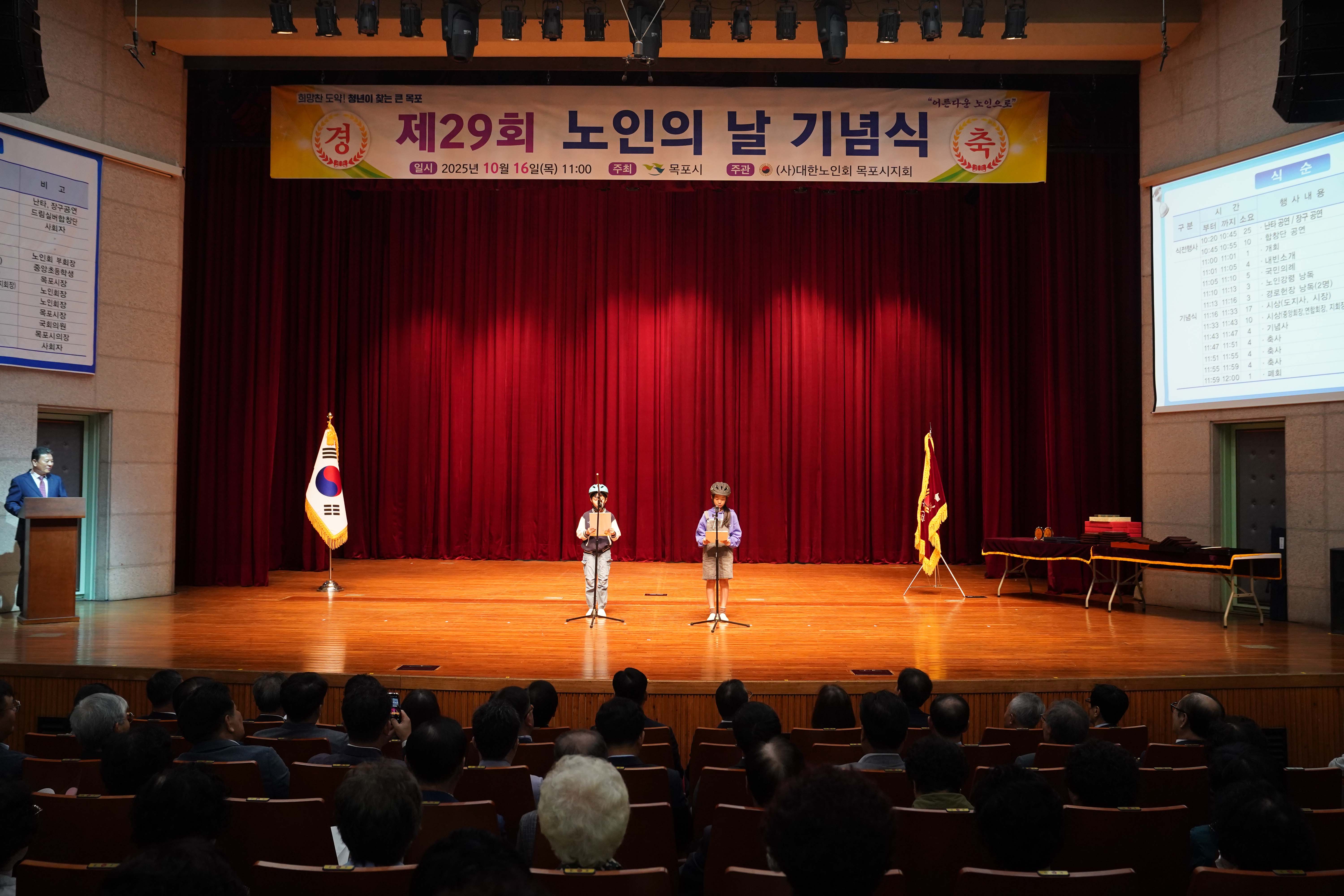 '제29회 노인의 날 기념식' 무대 중앙에서 두 어린이가 나란히 서서 발표를 진행하는 정면 모습. 무대 좌측에는 태극기가, 우측에는 단체기가 세워져 있음.