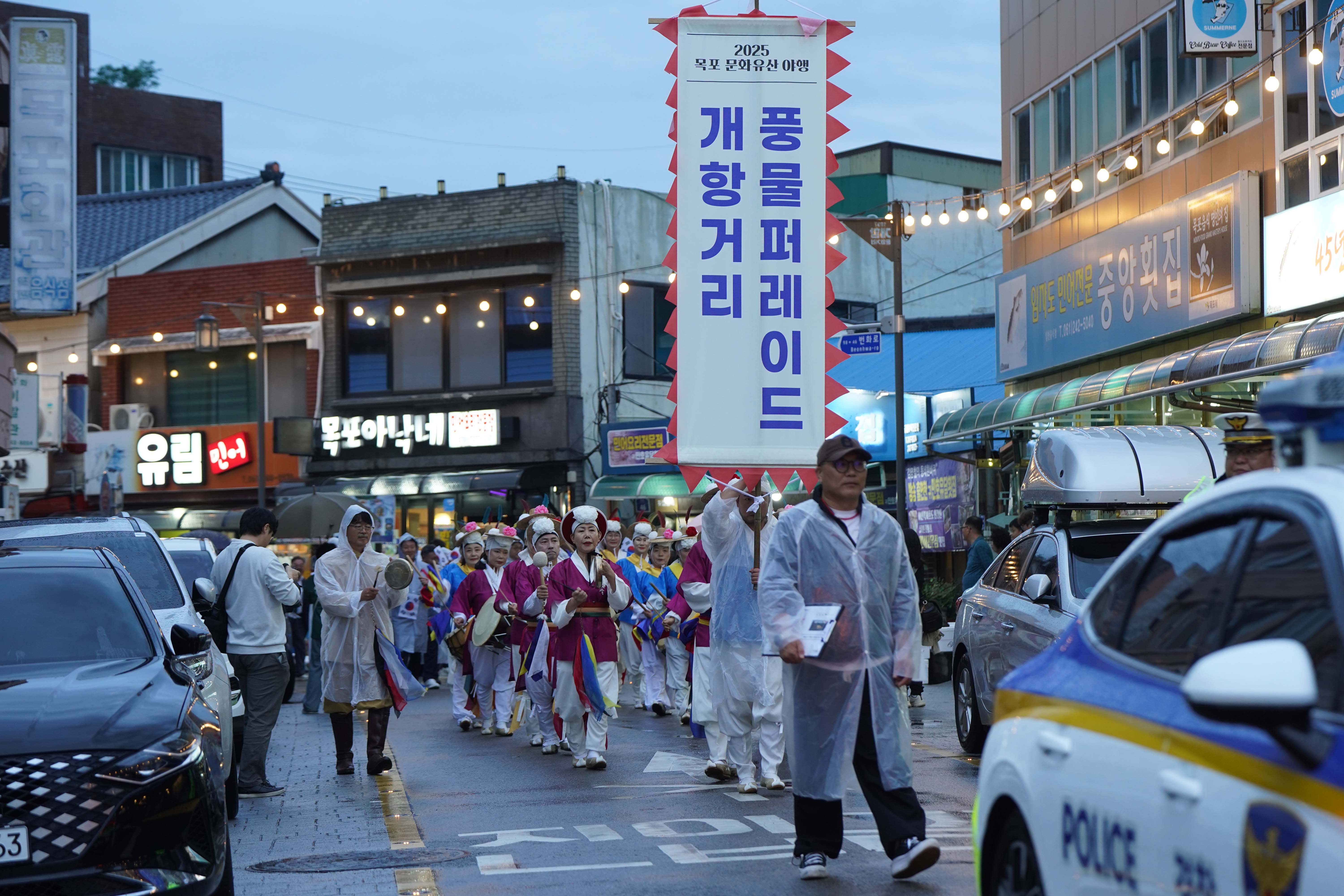 거리 행진의 전경. 상단에 '2025 목포 문화유산 야행'이라 적힌 대형 기치에 '풍물 퍼레이드 개항거리'라는 문구가 크게 쓰여 있음. 도로에는 경찰차와 우비를 입은 시민들이 보임.