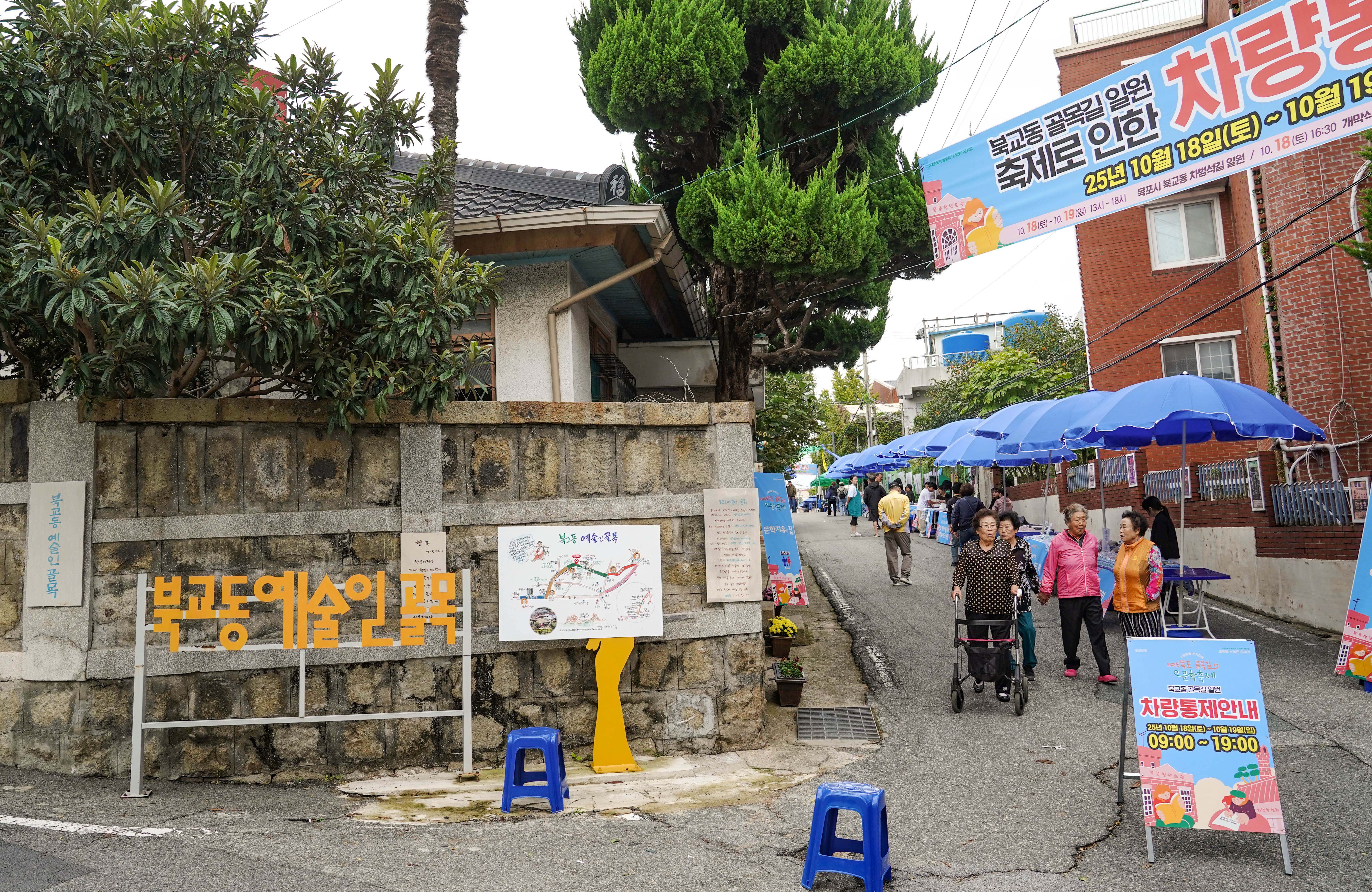 북교동 예술인 골목 입구 전경. '북교동 골목길 일원 축제로 인한 차량통제' 안내 현수막(10월 18일~19일)과 차량통제 안내판이 세워져 있으며, 골목을 따라 파란색 파라솔 부스들이 줄지어 있음.