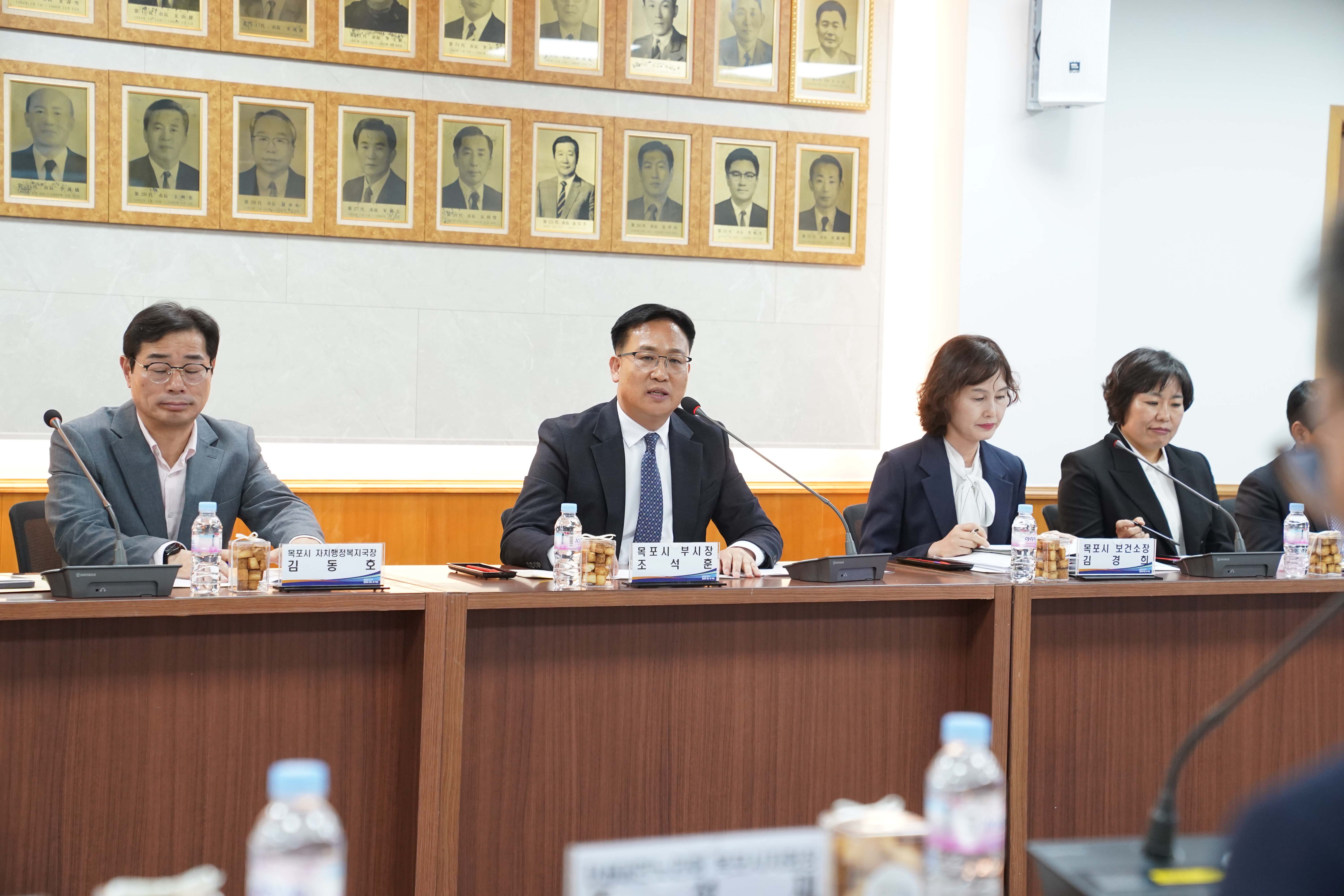 회의실 상단에 역대 시장들의 사진 액자가 걸려 있고, 테이블 중앙에 '목포시 부시장 조석훈' 명패가 놓여 있음. 조석훈 부시장이 마이크 앞에서 발언하고 있으며 좌측에는 '목포시 자치행정복지국장 김동호', 우측에는 '목포시 보건소장 김경희' 명패가 보임.