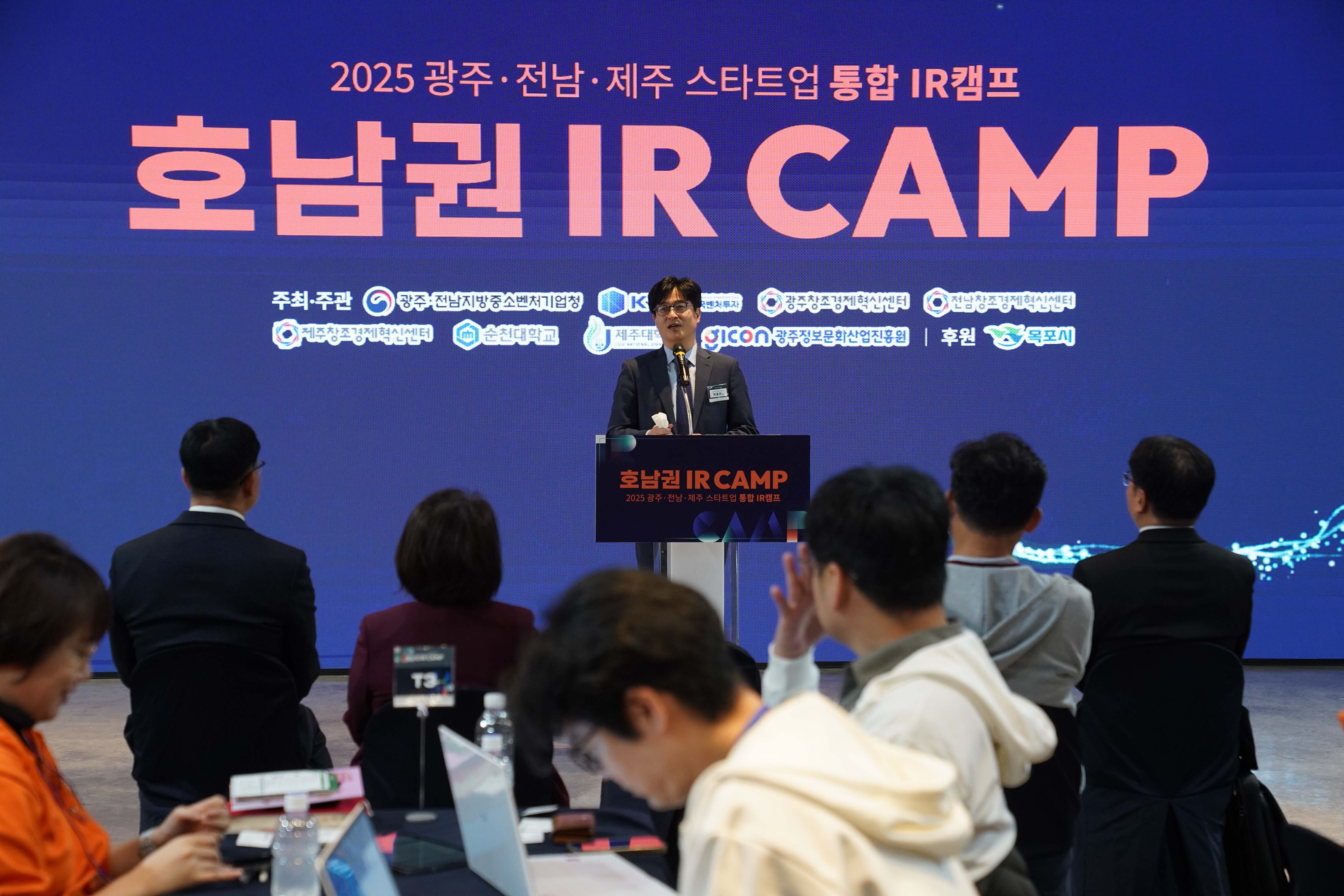 '2025 광주·전남·제주 스타트업 통합 IR캠프 호남권 IR CAMP' 행사장 무대에서 관계자가 인사말을 하는 모습. 배경 스크린에 광주·전남지방중소벤처기업청, KVIC, 광주/전남/제주창조경제혁신센터 등 주최·주관사와 후원 목포시 로고가 보임.