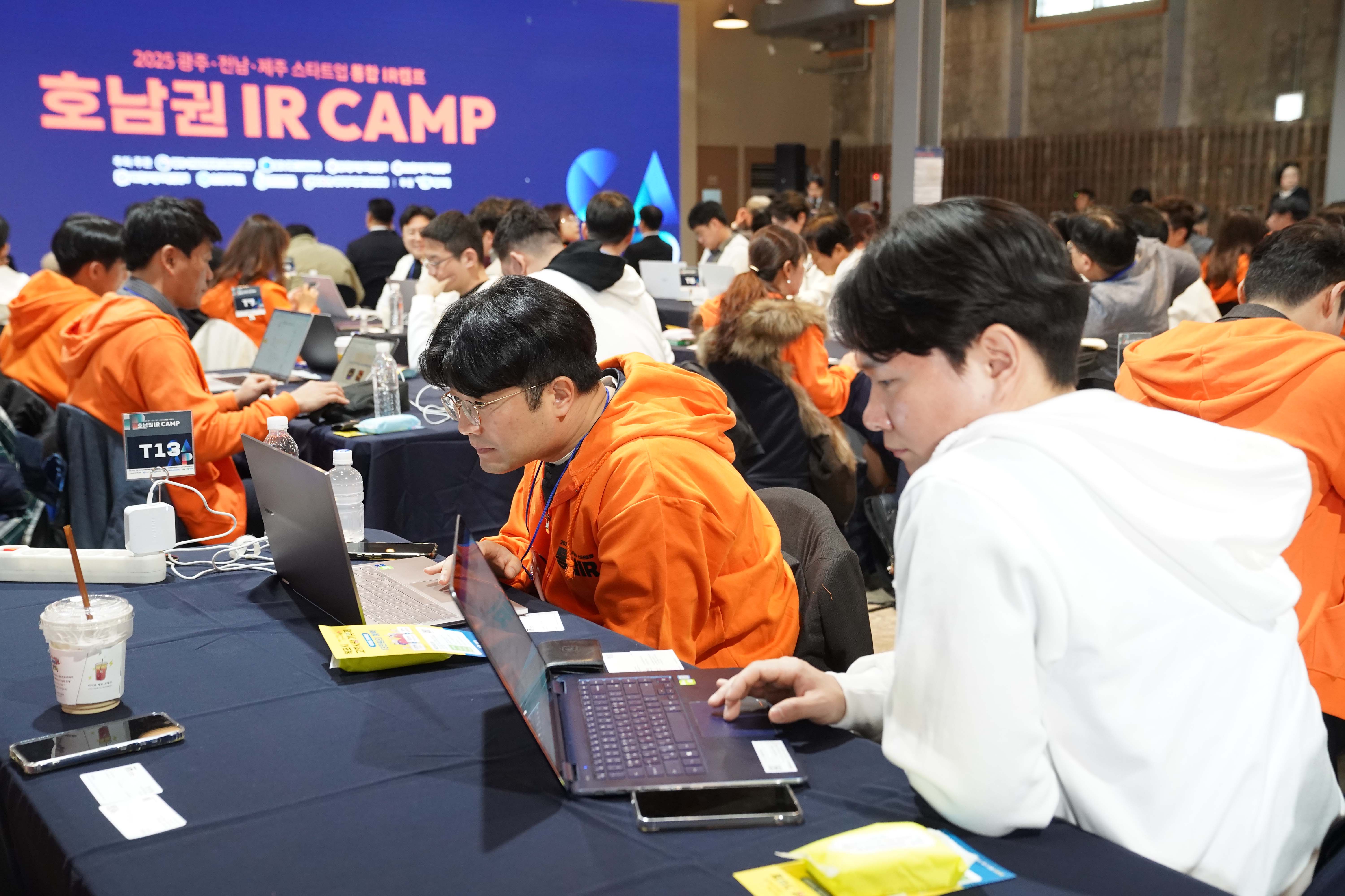 '호남권 IR CAMP' 행사장 내부 전경. 주황색과 흰색 후드티를 입은 스타트업 관계자들이 노트북 앞에 앉아 집중하며 업무를 보거나 토론하고 있는 모습.