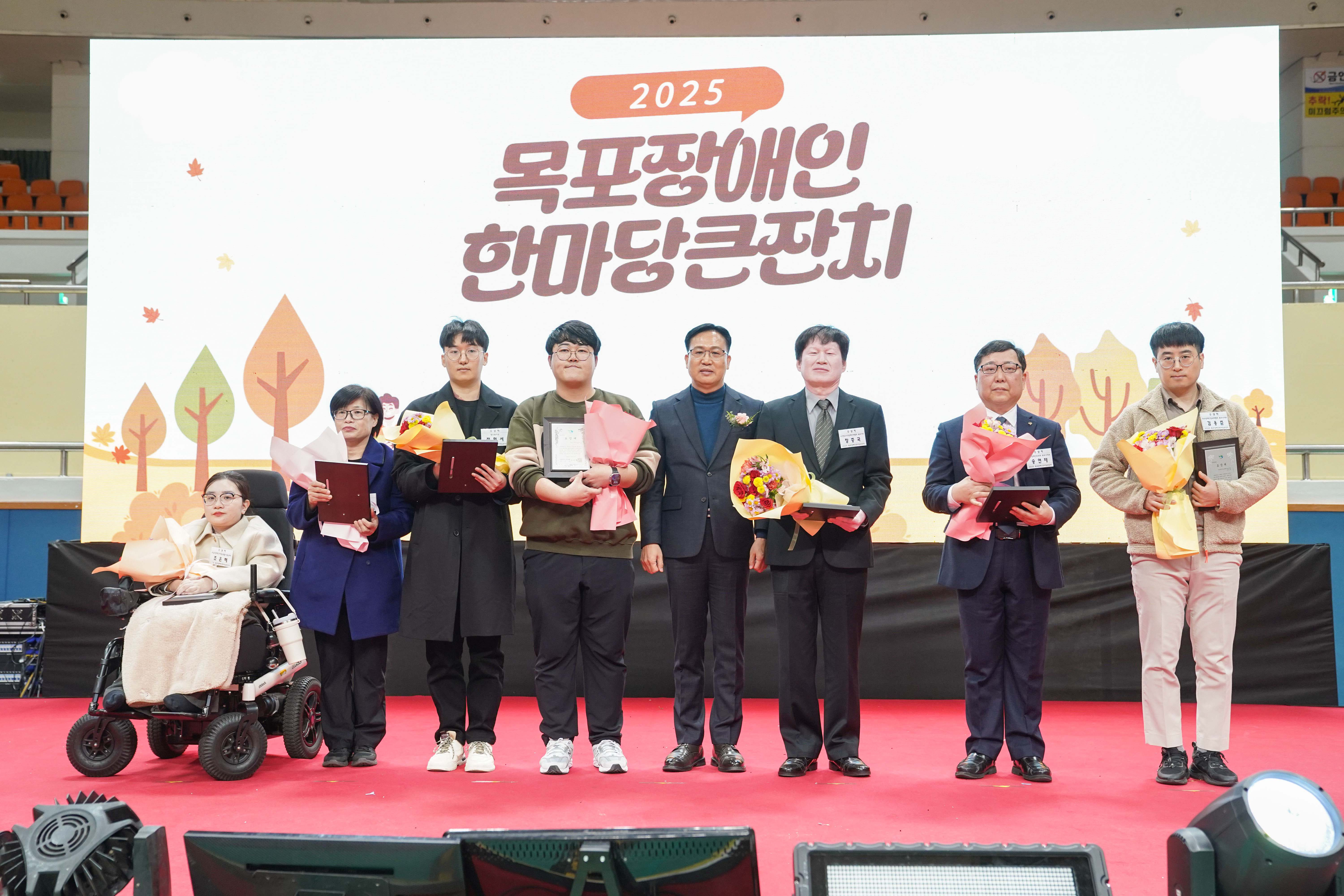 '2025 목포장애인 한마당큰잔치'라고 적힌 대형 스크린 무대 위에서 조석훈 부시장과 수상자들이 상장과 꽃다발을 들고 기념 촬영을 하는 모습. 휠체어를 탄 수상자를 포함해 총 7명이 나란히 서 있음.