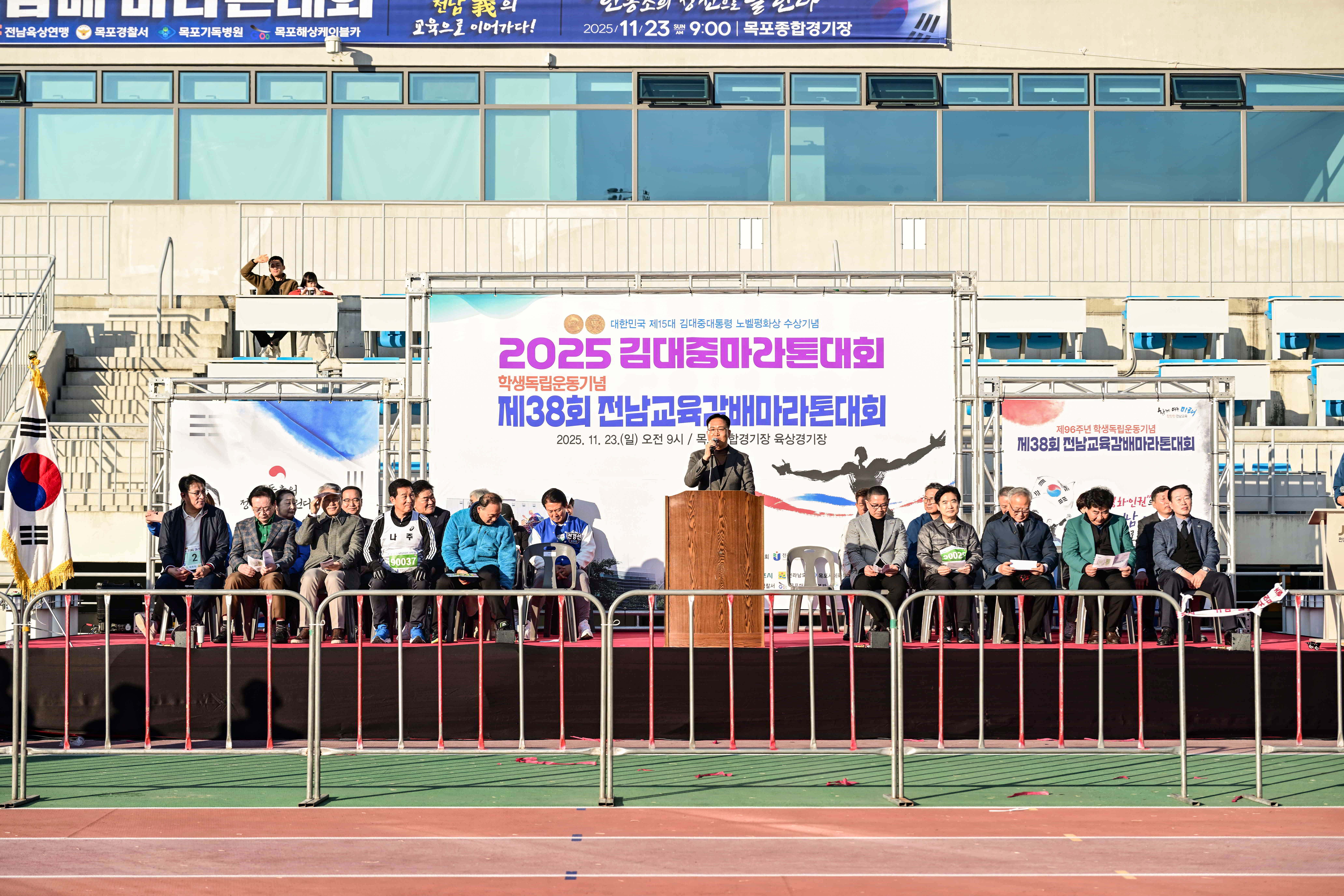 목포종합경기장 육상경기장에 마련된 마라톤 대회 메인 무대 전경. 조석훈 부시장이 연단에서 축사를 하고 있으며, 양옆으로 내빈들이 배석해 있음. 무대 앞에는 안전 펜스가 설치되어 있고 경기장 트랙이 보임.