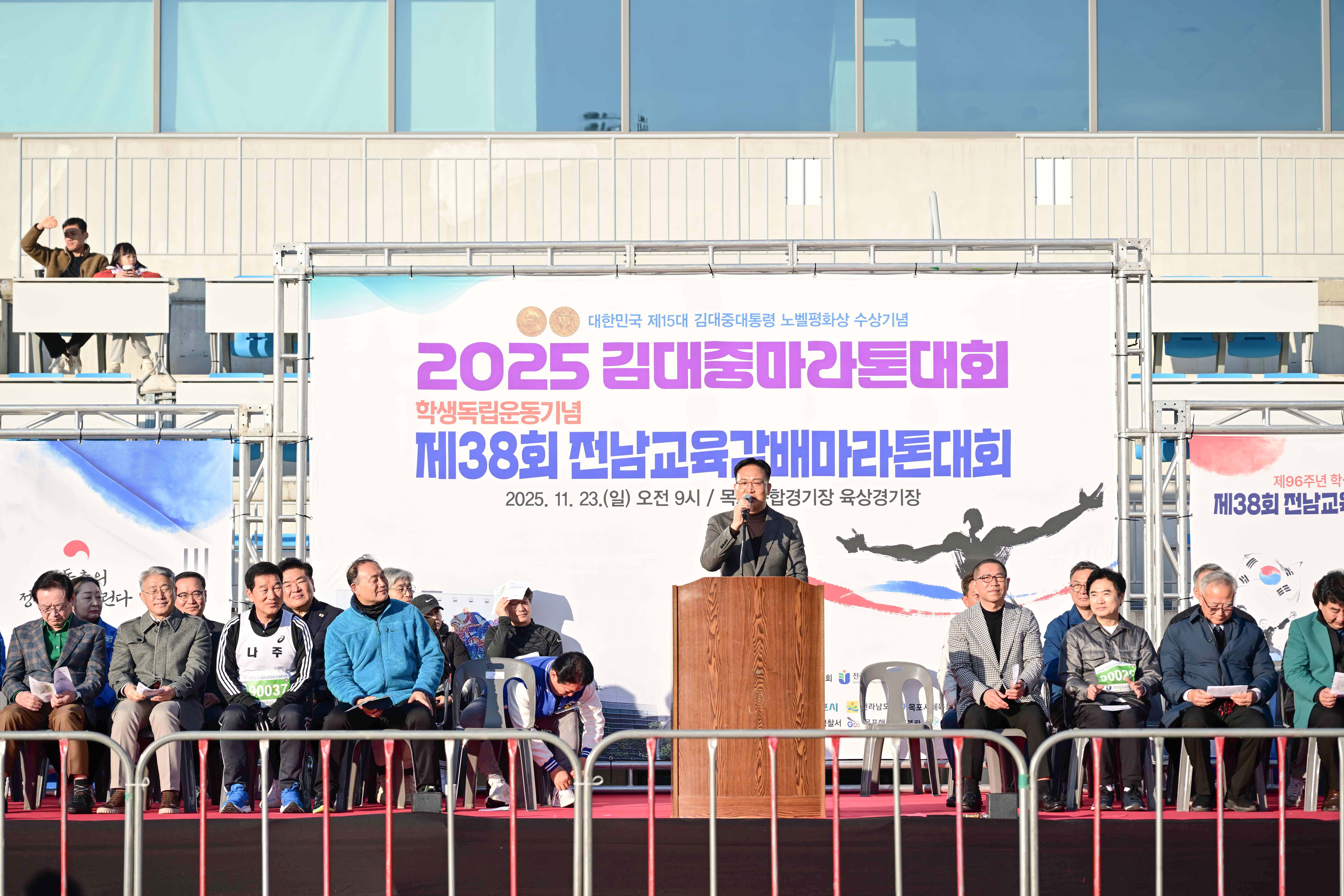 마라톤 대회 무대 위에서 마이크를 잡고 발언 중인 조석훈 부시장. 배경에는 '2025. 11. 23.(일) 오전 9시 / 목포종합경기장 육상경기장'이라는 대회 일시와 장소 정보가 포함된 대형 스크린이 보임.